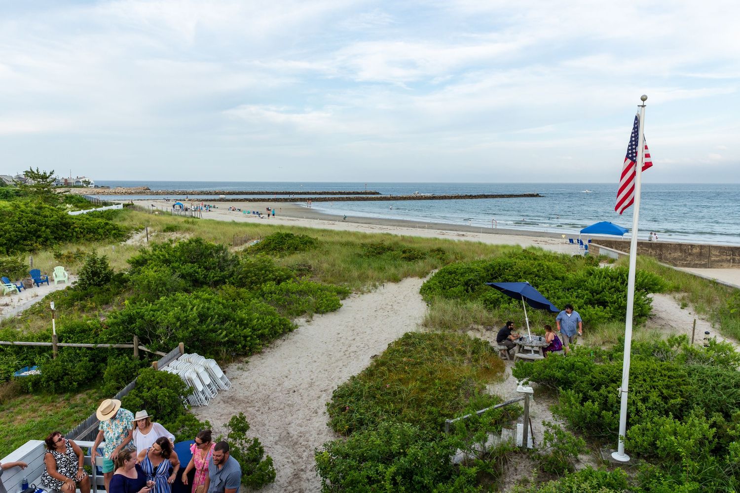 A scenic coastal pathway leads to a beach with an American flag flying beside the Atlantic Ocean on a sunny day.
