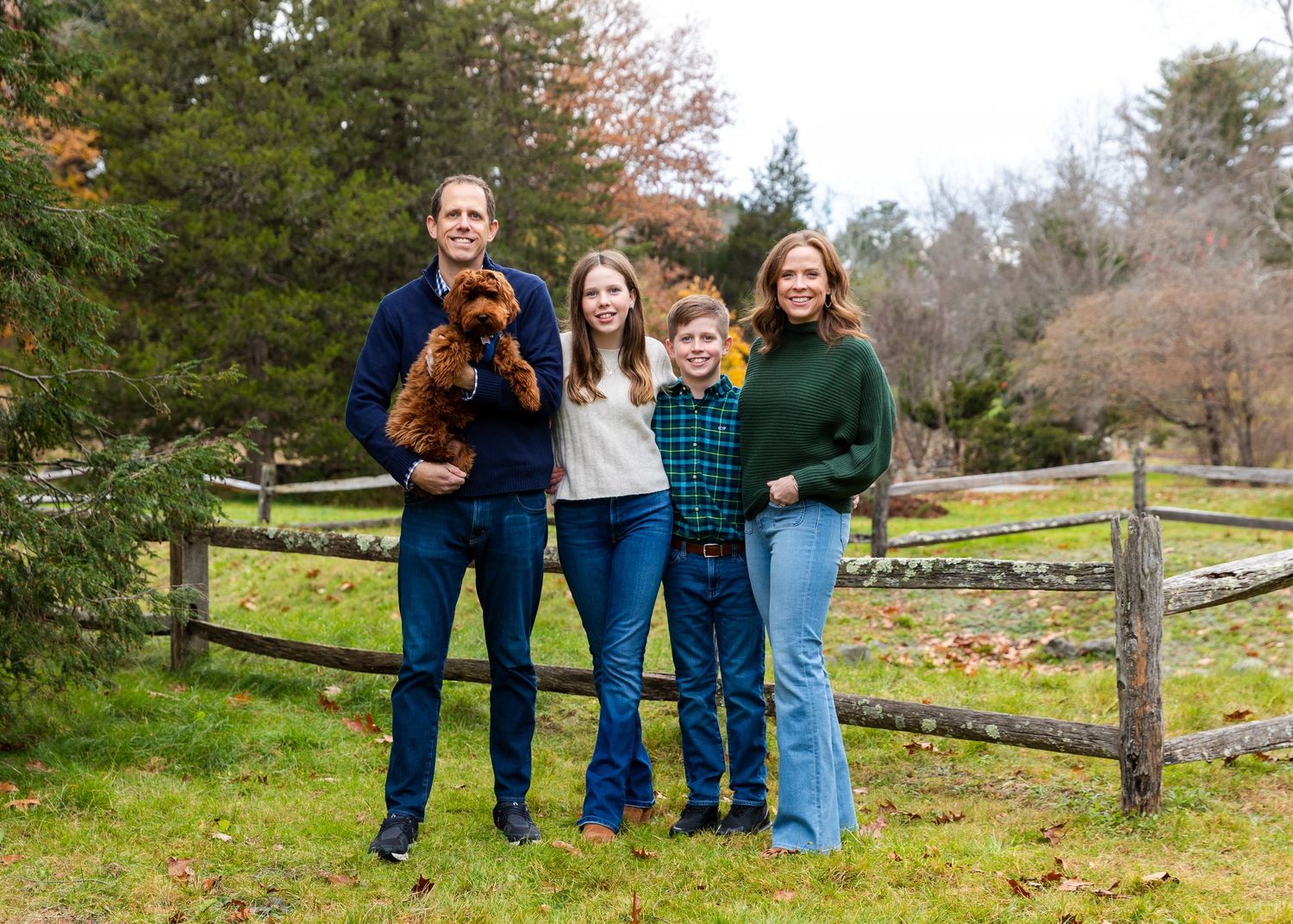 Family poses together with their dog in front of a rustic wooden fence on an autumn day.