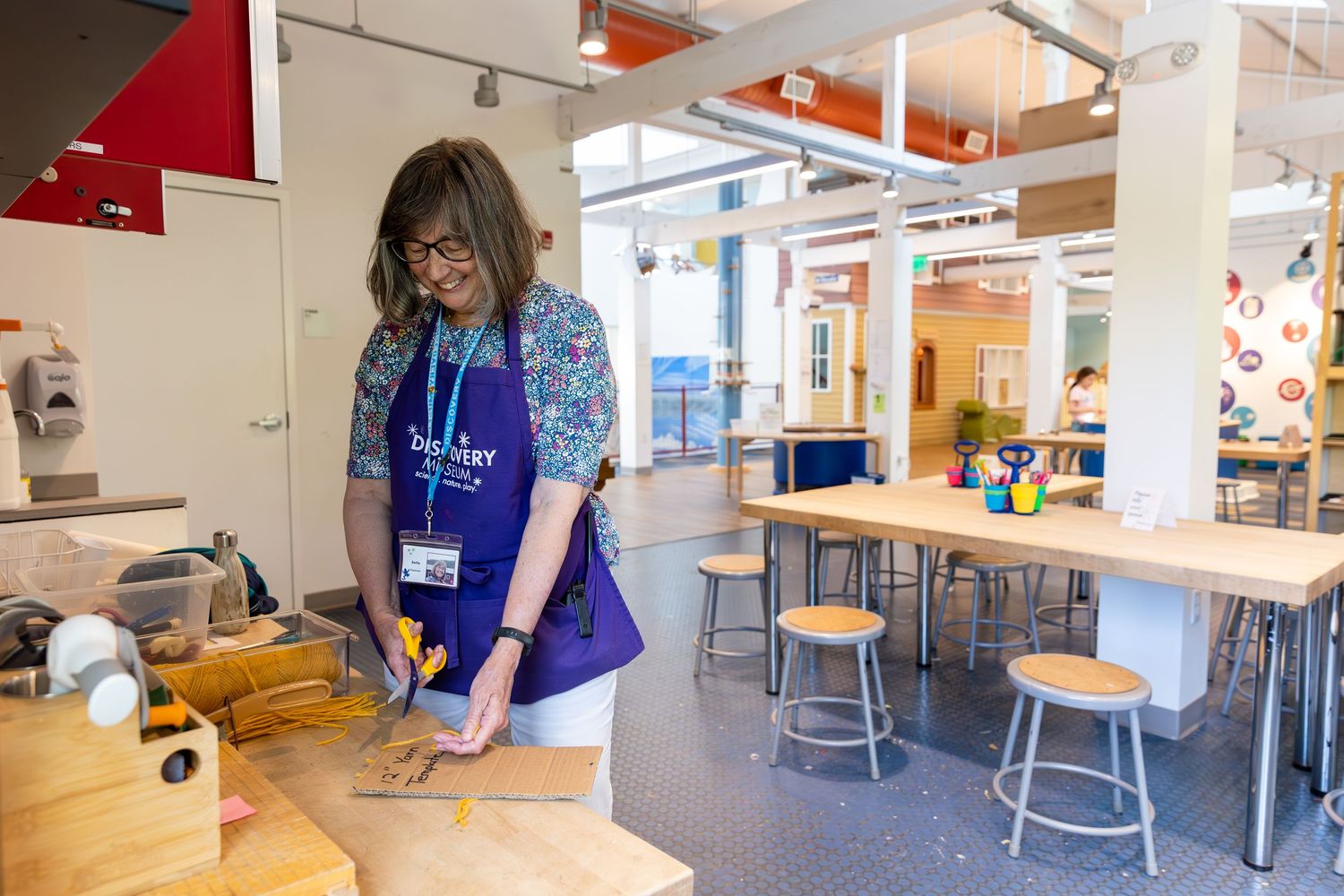 Someone in a blue apron and name badge works at a butcher block counter in a bright modern cafeteria setting.