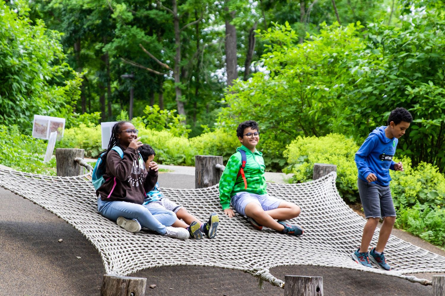Three people sitting on an outdoor hammock net structure in a lush green park setting.
