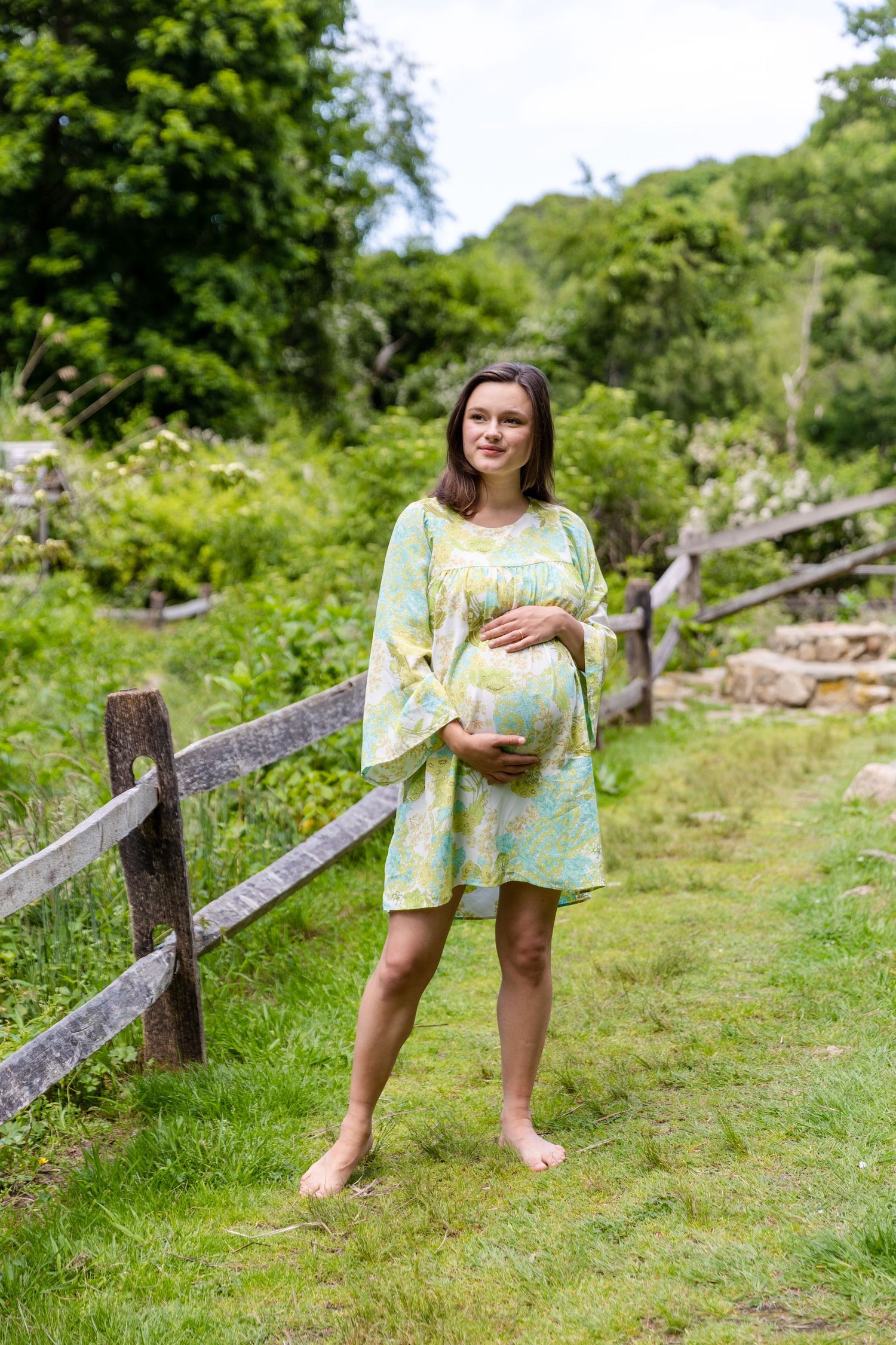 Woman in floral summer dress posing by rustic wooden fence in lush green countryside setting.