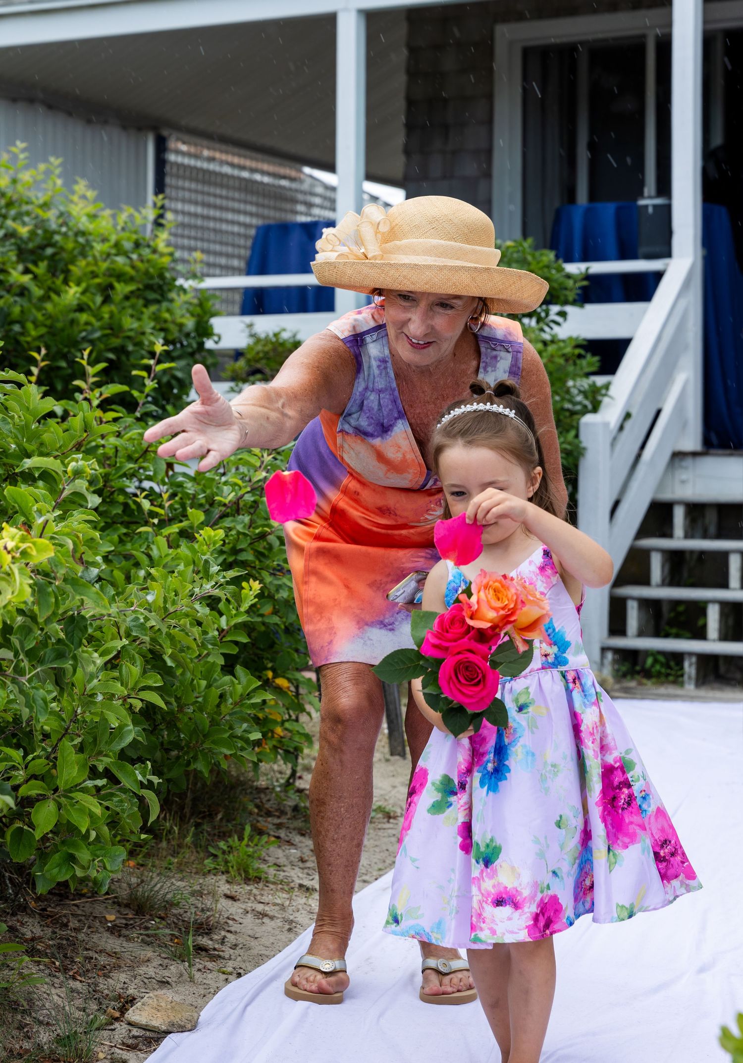 Two people in colorful summer dresses stand together outdoors by garden steps with bright flowers.