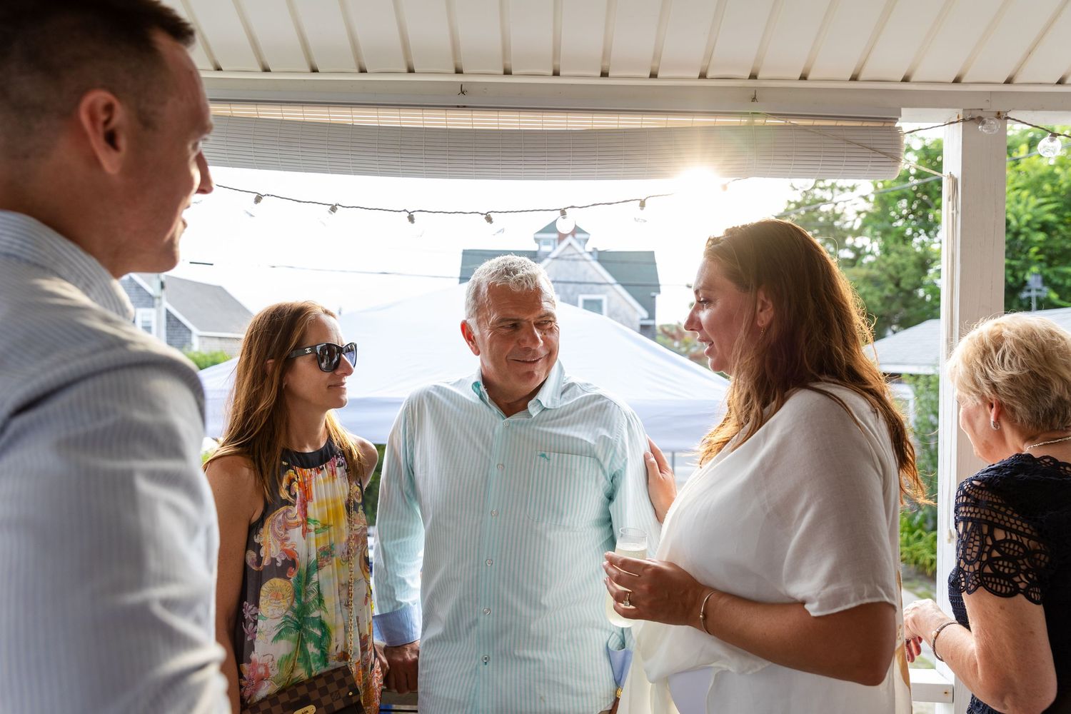 A small group of people gather and converse on a covered porch with string lights during a social gathering.