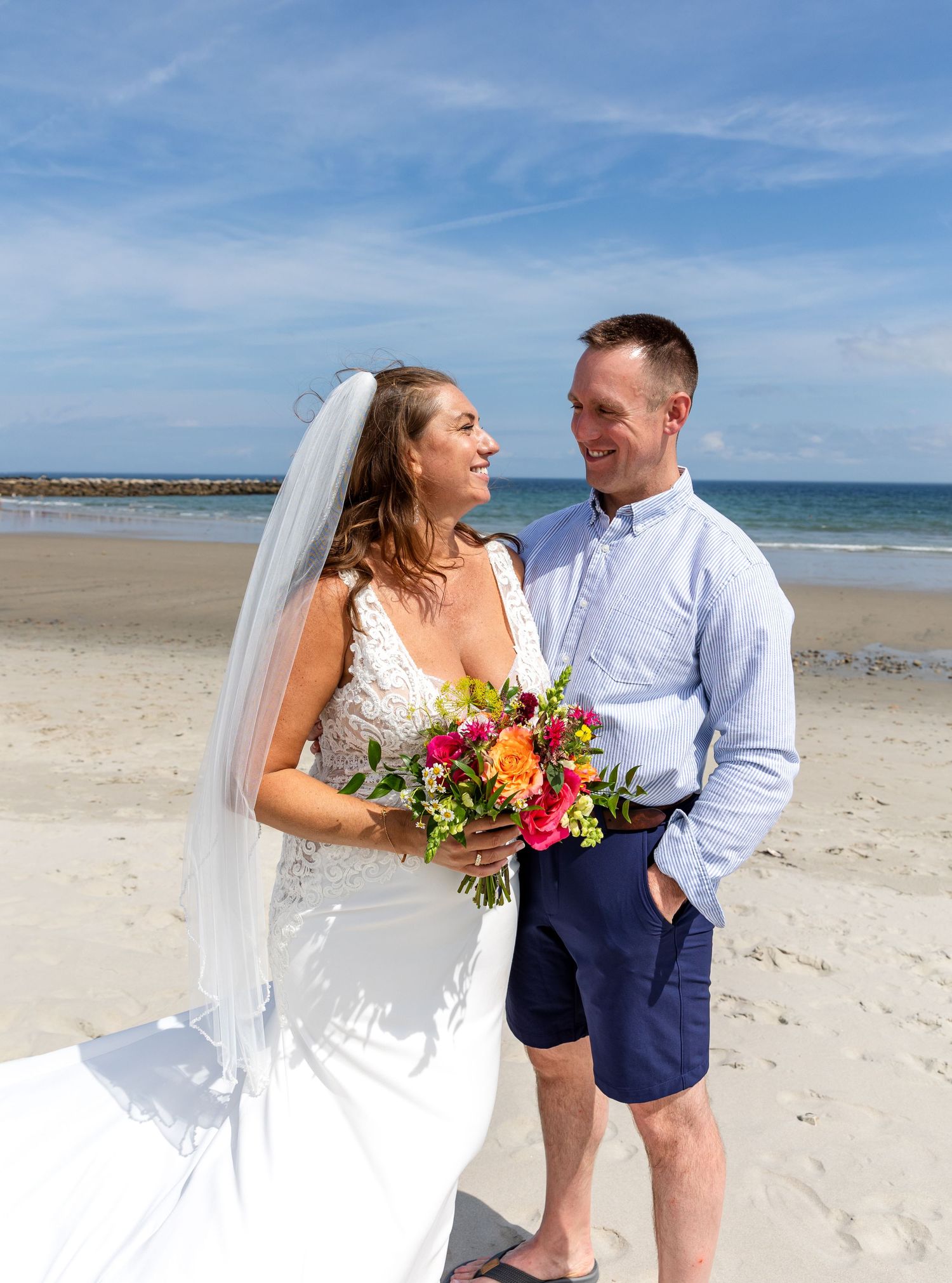 A joyful beach wedding ceremony with a bride in white dress holding colorful bouquet on a sunny day by the ocean.