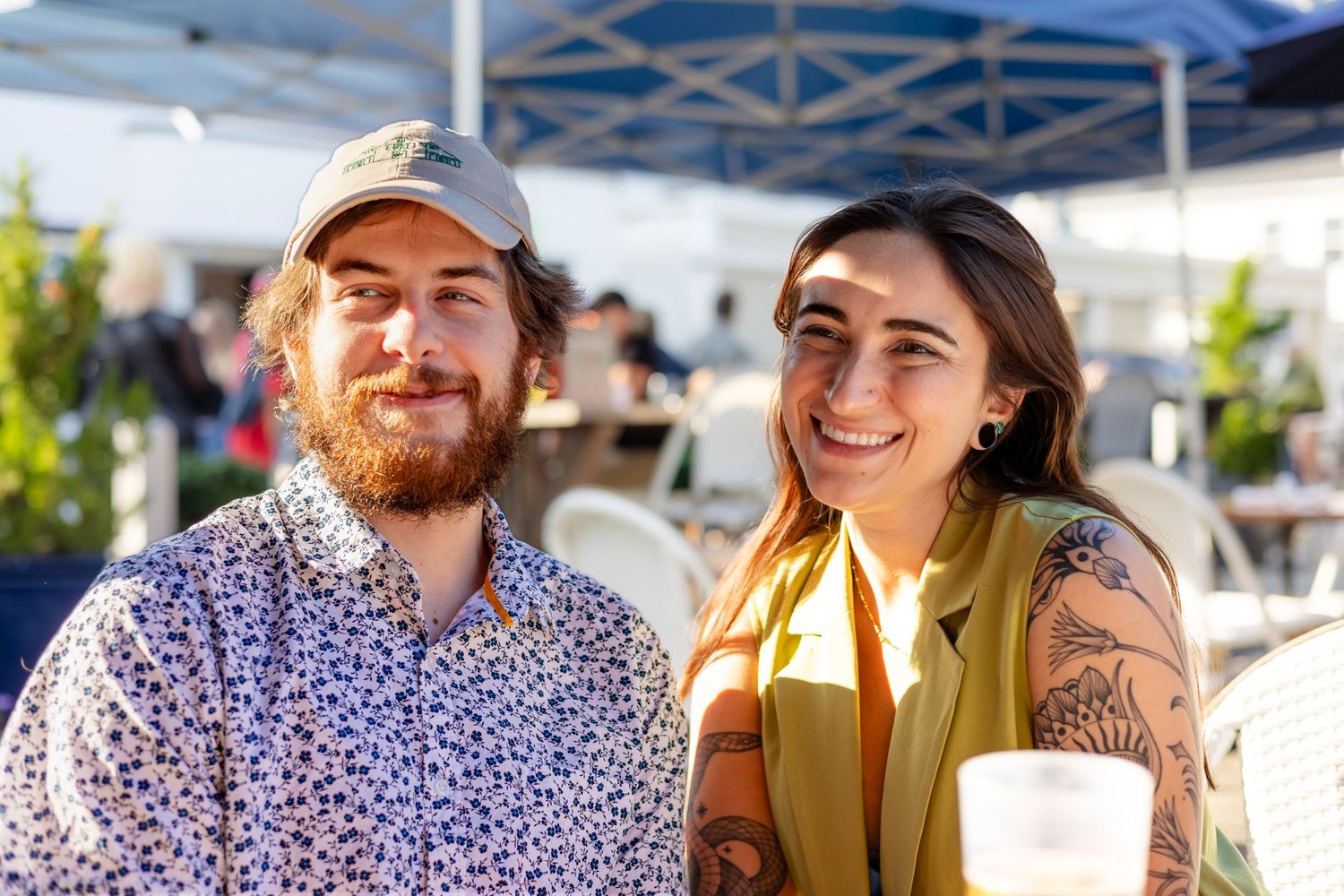 Two people smile at an outdoor market under a blue tent on a sunny day.