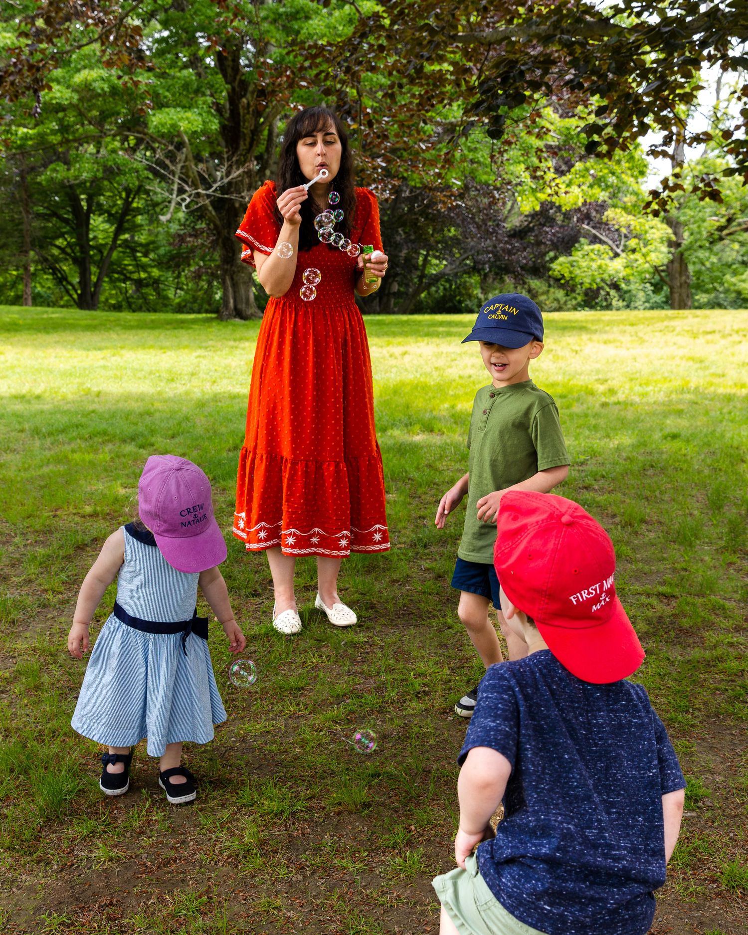 Group of children playing and blowing bubbles in a lush green yard with a woman in a red dress watching.