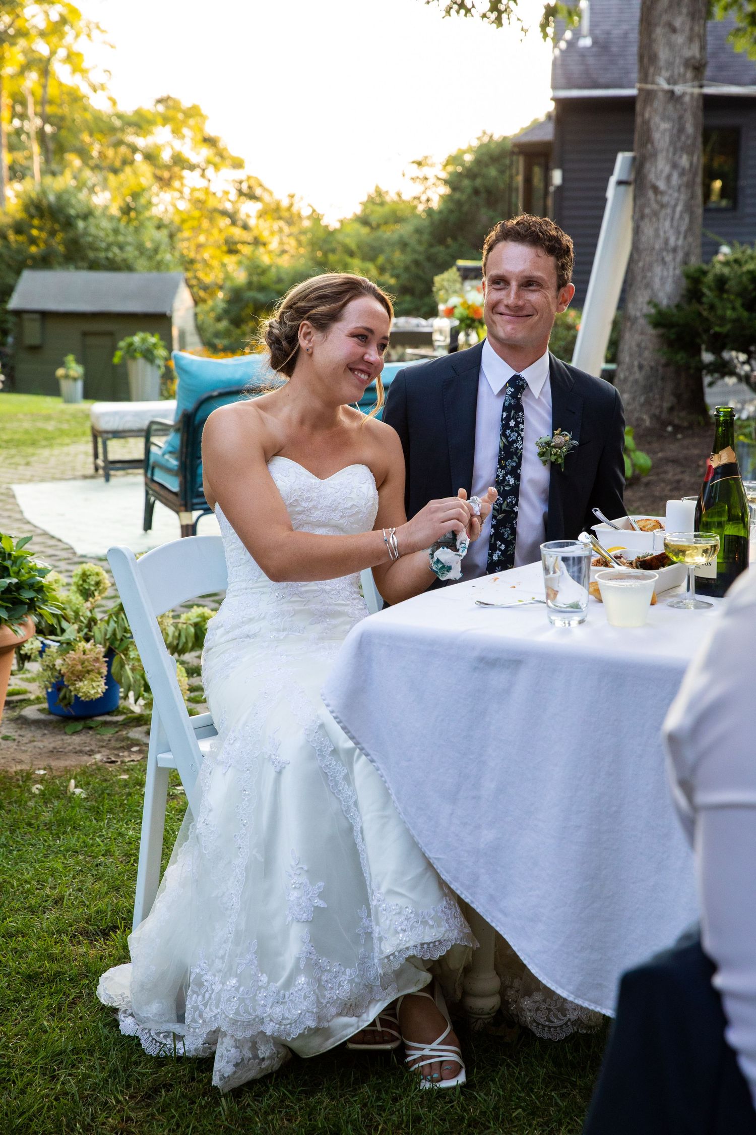 A happy couple enjoys their outdoor wedding dinner at a table with white linens during golden hour.