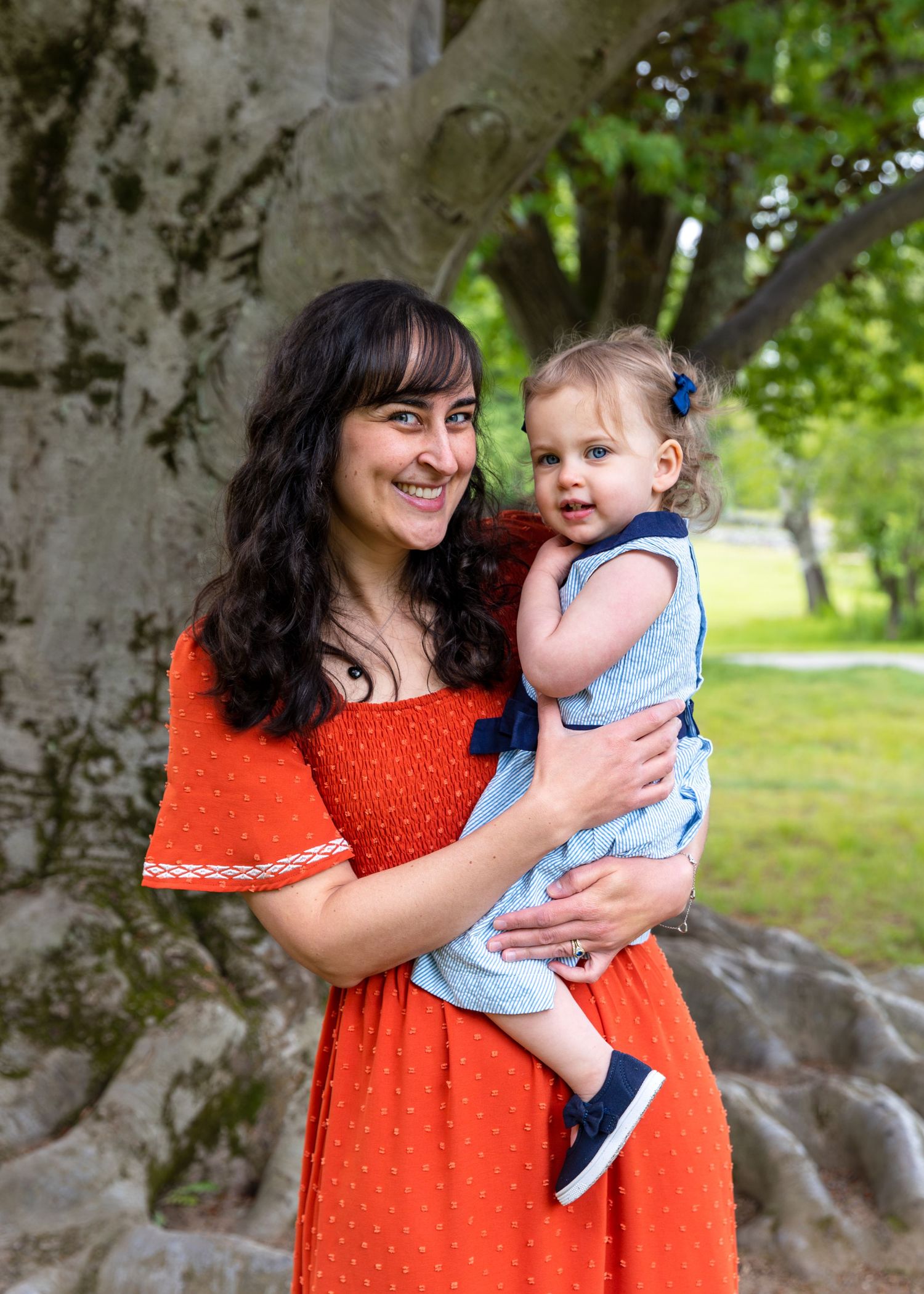 A woman in a bright coral dress embraces a baby wearing sailor-style clothing while standing in front of large tree trunks.