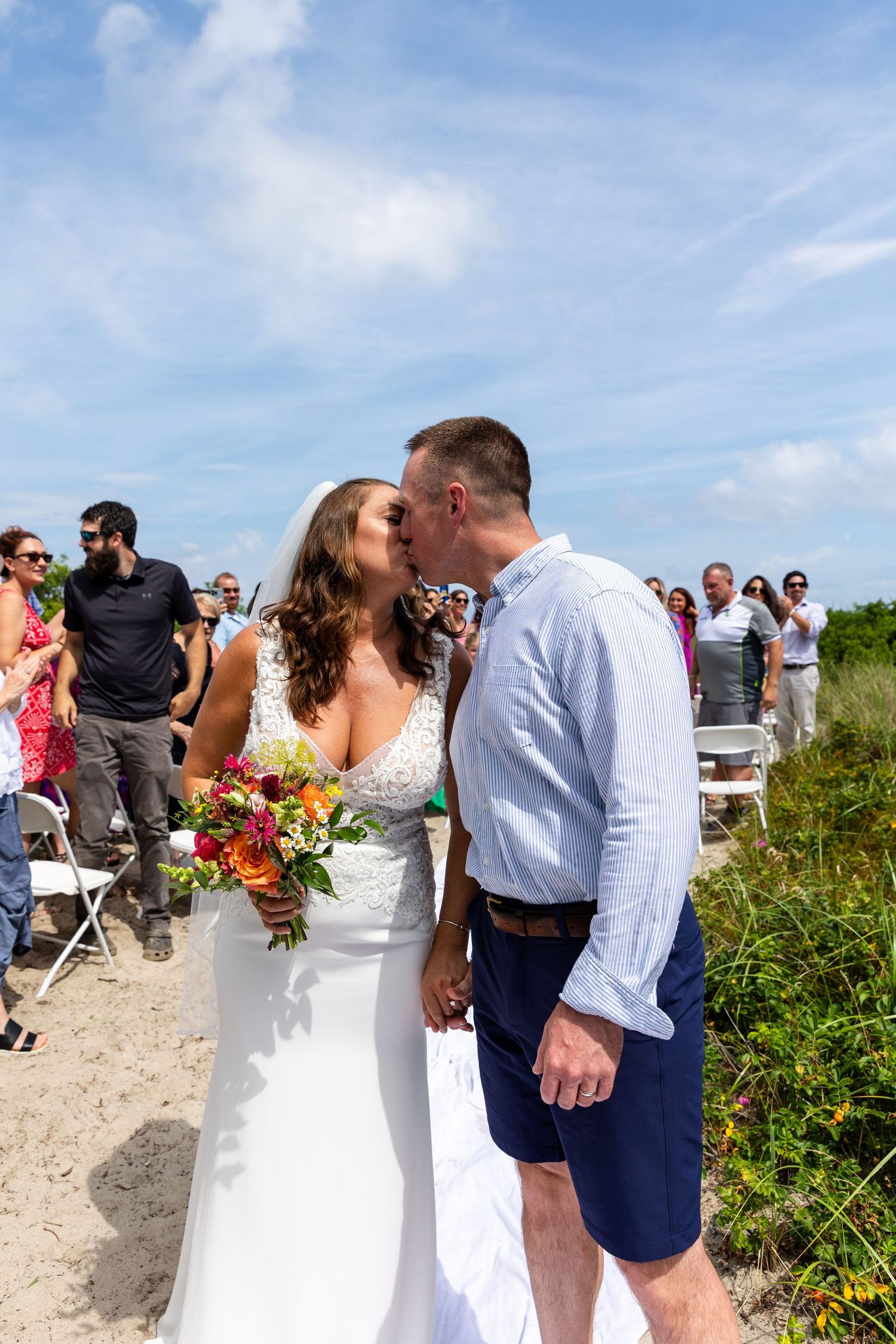 A seaside wedding celebration takes place on a sandy beach path with guests seated on white chairs under blue skies.