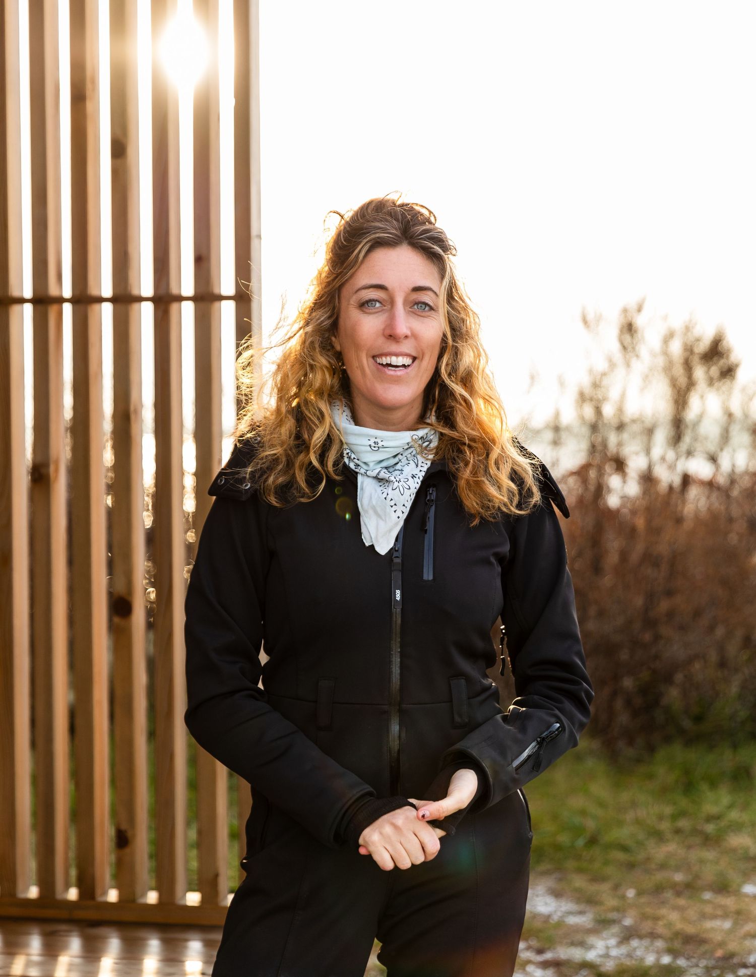 Woman wearing black jacket and white bandana smiling outdoors in front of wooden slats during golden hour lighting.