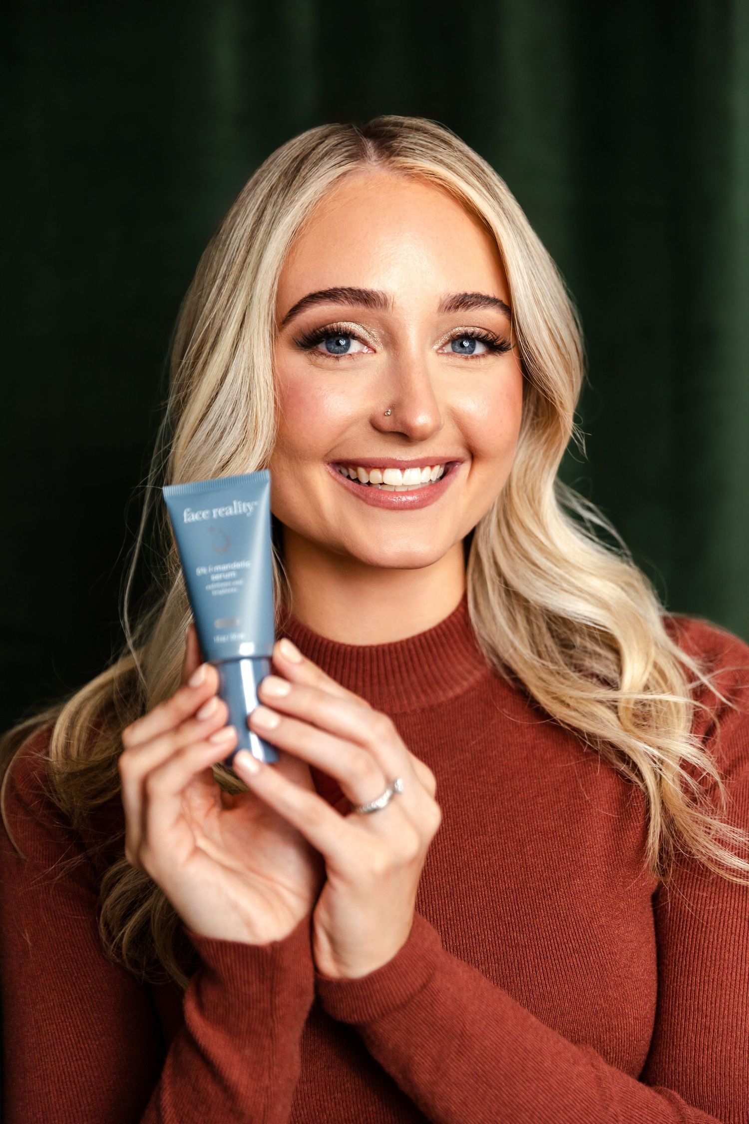 Woman in rust-colored sweater smiles while holding blue skincare product against green background.
