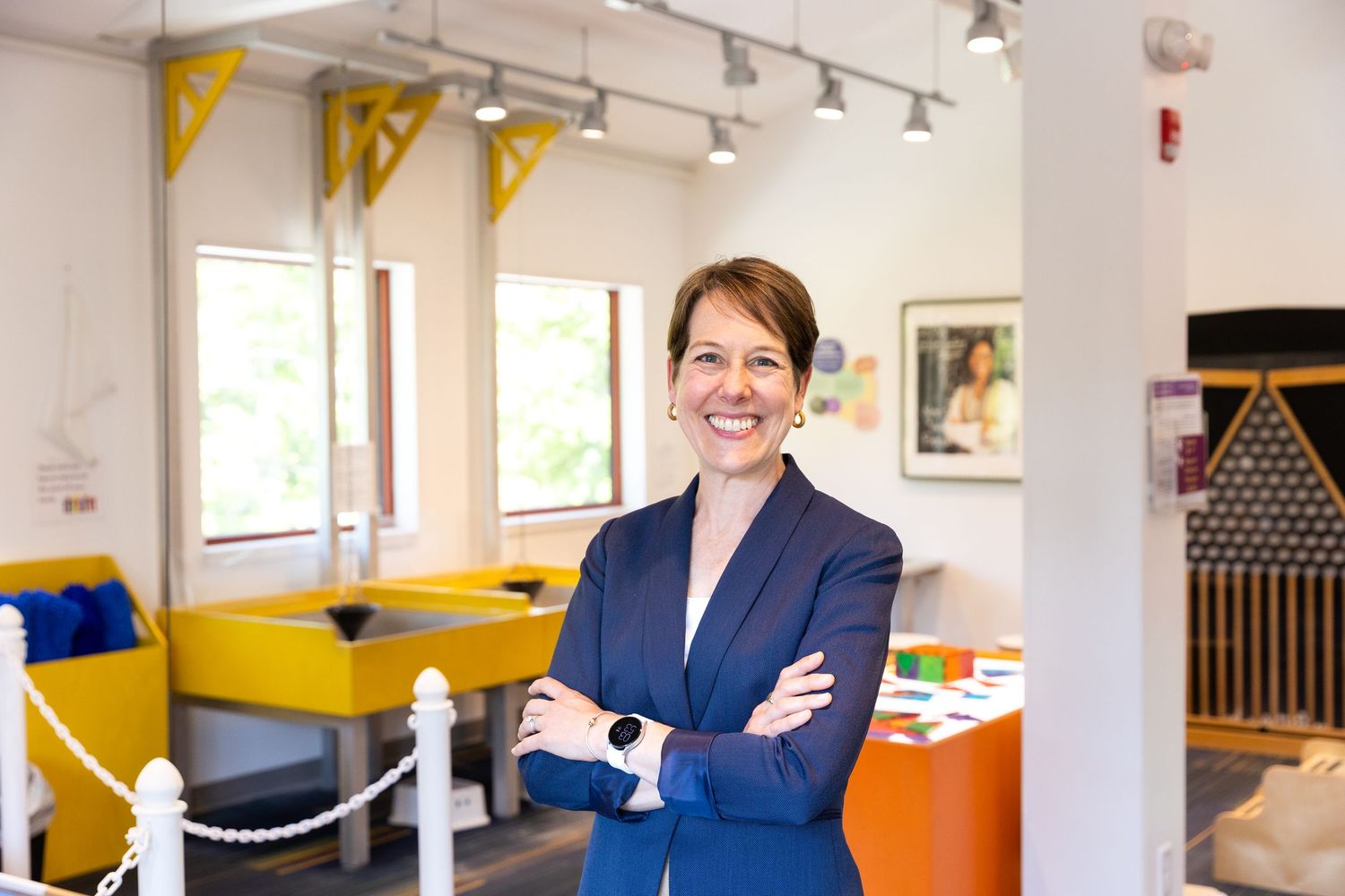 Professional wearing navy blazer stands confidently with arms crossed in modern office with yellow accents.
