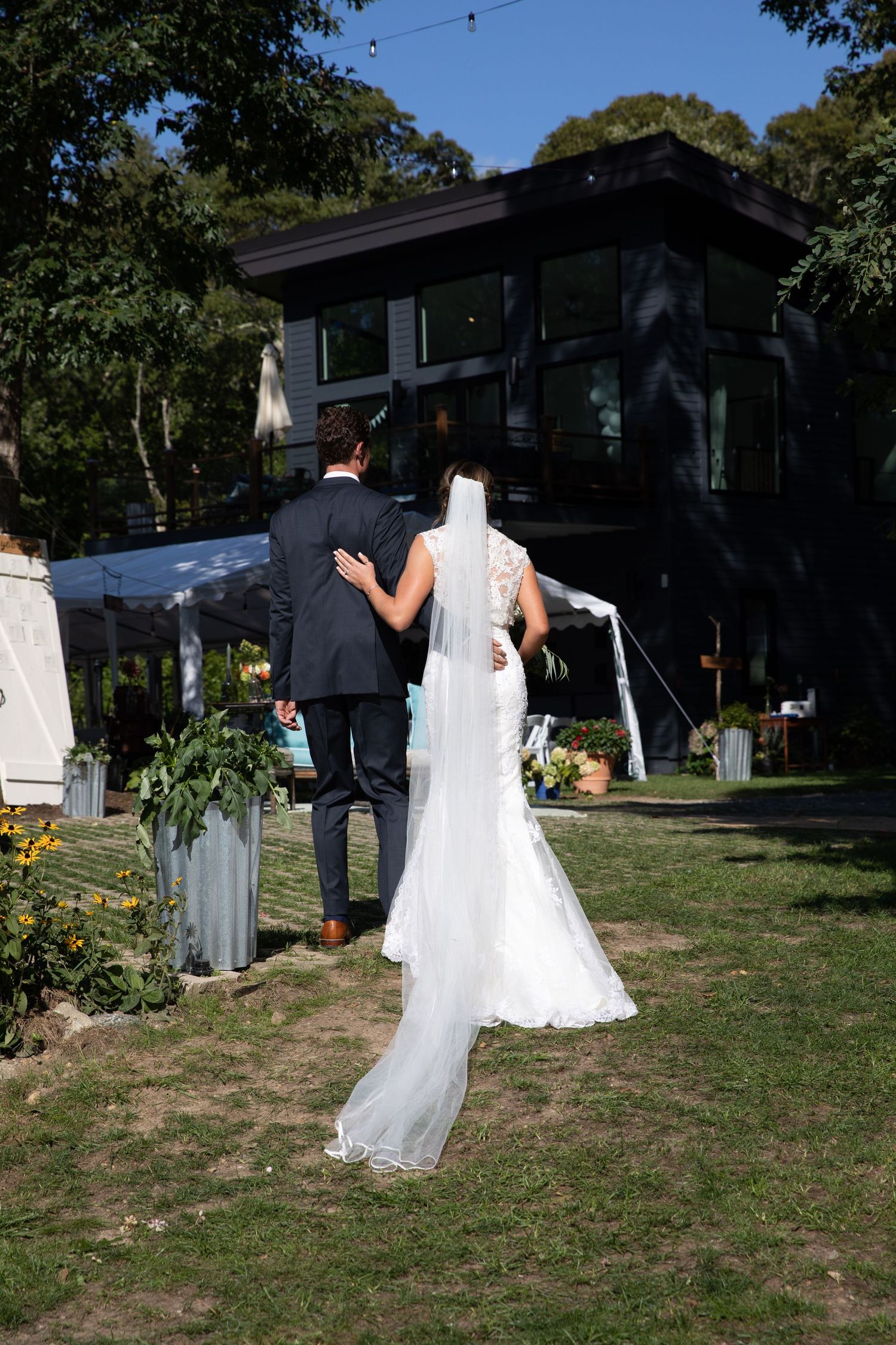 A couple in wedding attire stands on a lawn during an outdoor ceremony with a dark house in the background.