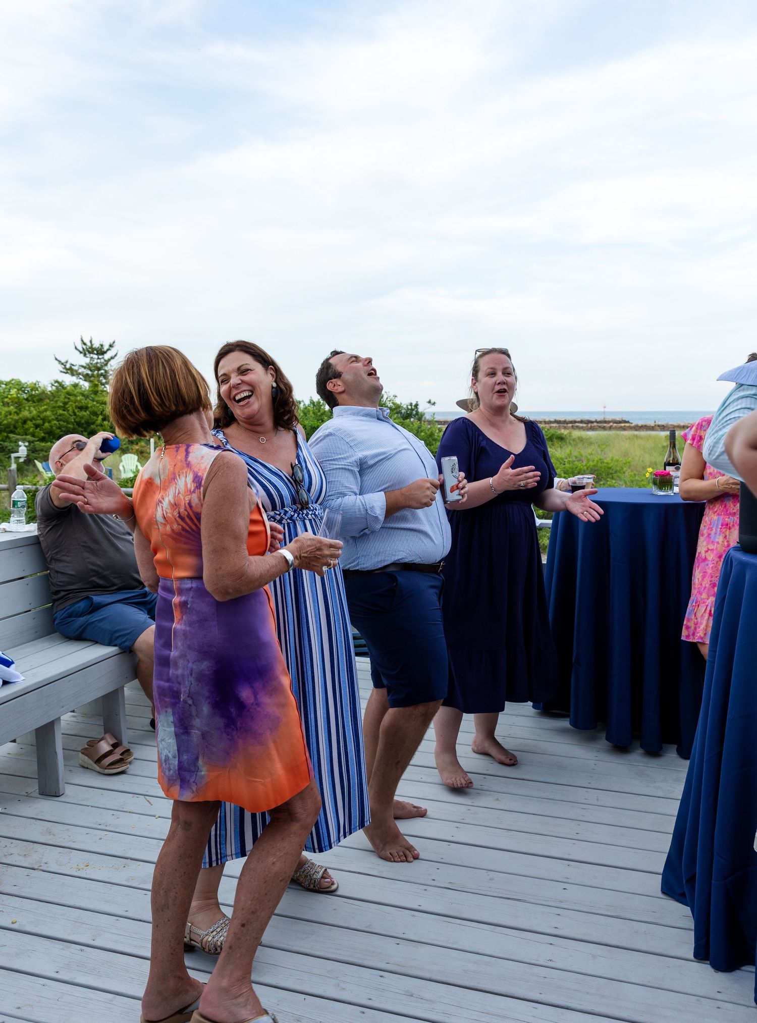 People enjoying themselves while dancing and laughing at an outdoor celebration on a wooden deck with ocean view.