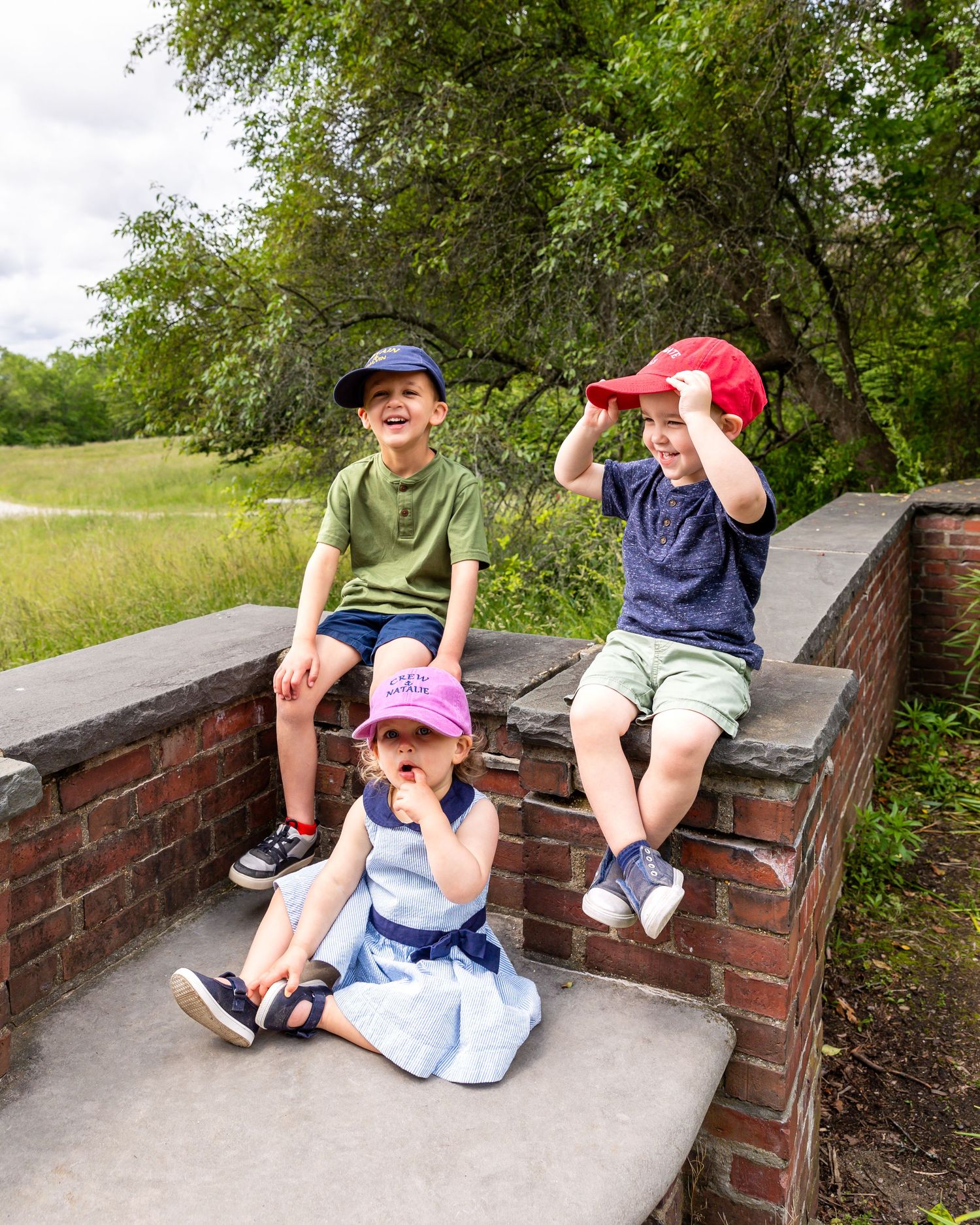 Children in colorful caps sit casually on a brick wall along a park path surrounded by green trees.