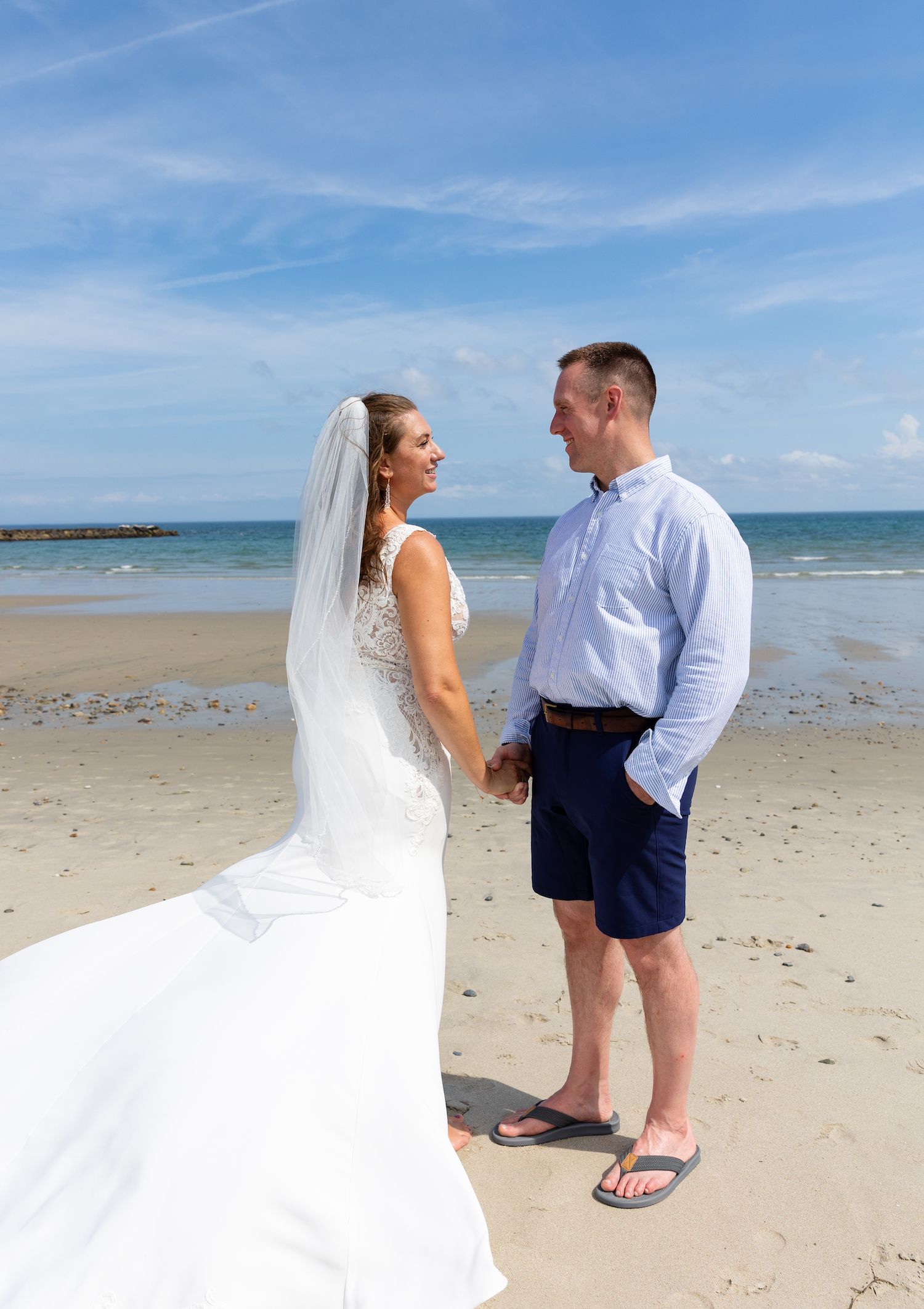 A scenic beach wedding scene with a couple standing on the sand, the groom wearing shorts and the bride in a flowing white gown.