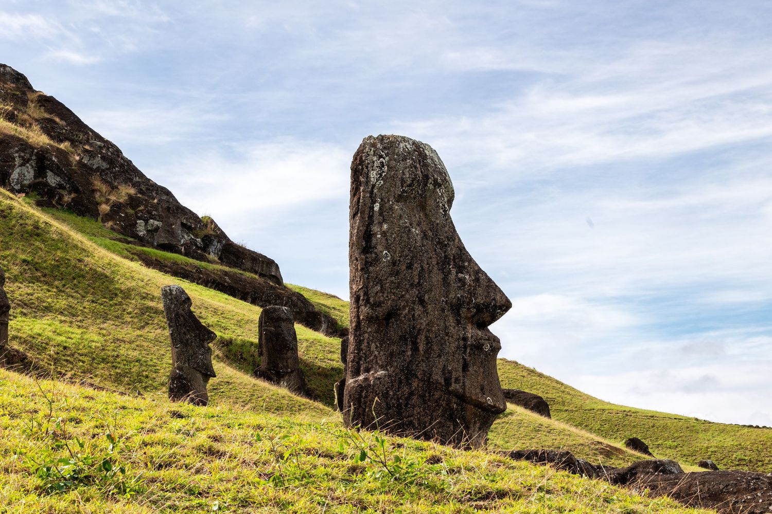 Ancient Moai stone statue stands on grassy hillside at Rano Raraku, Easter Island against blue sky.
