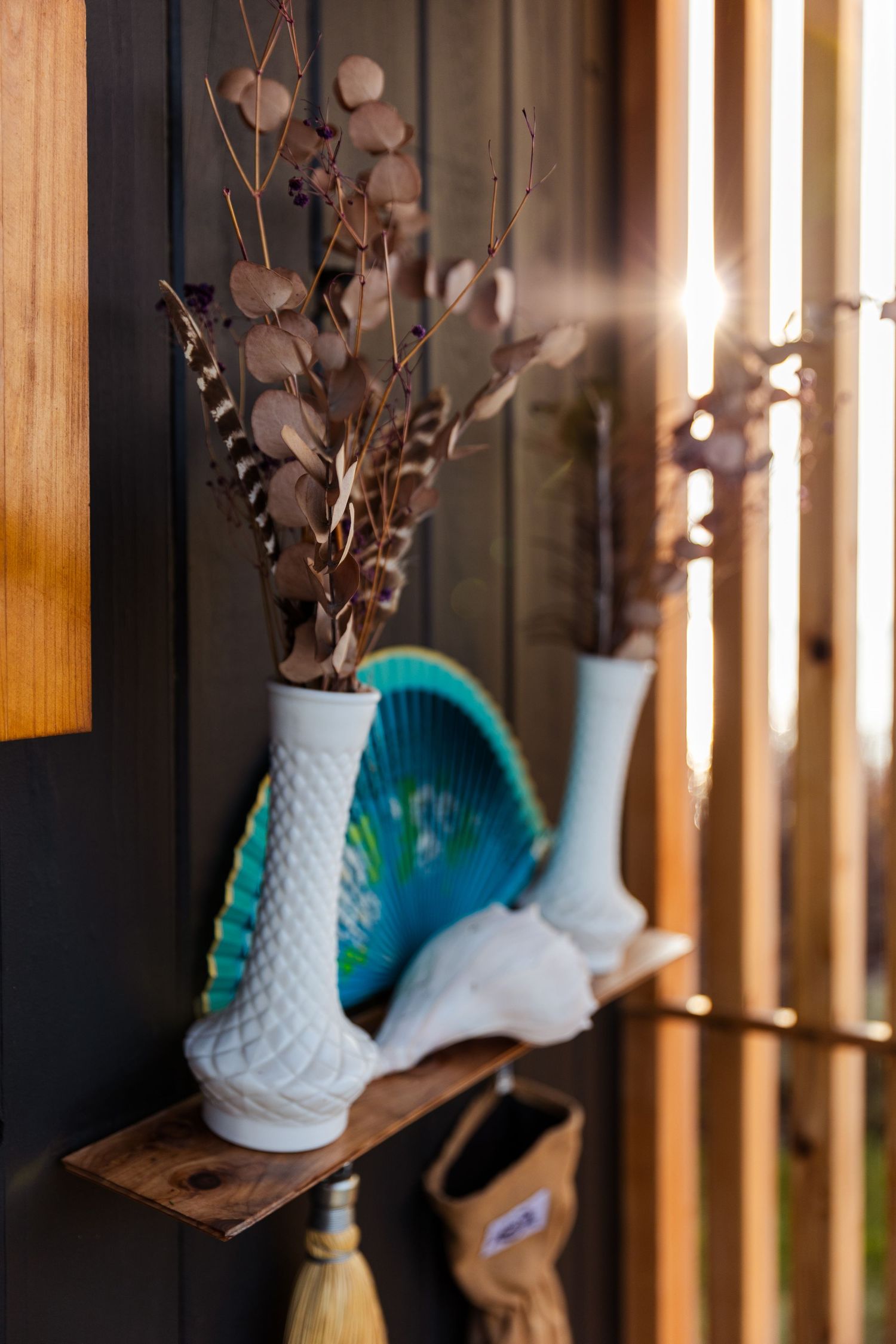 Milk glass vases with dried flowers sit on a wooden shelf beside a turquoise decorative piece, backlit by window light.