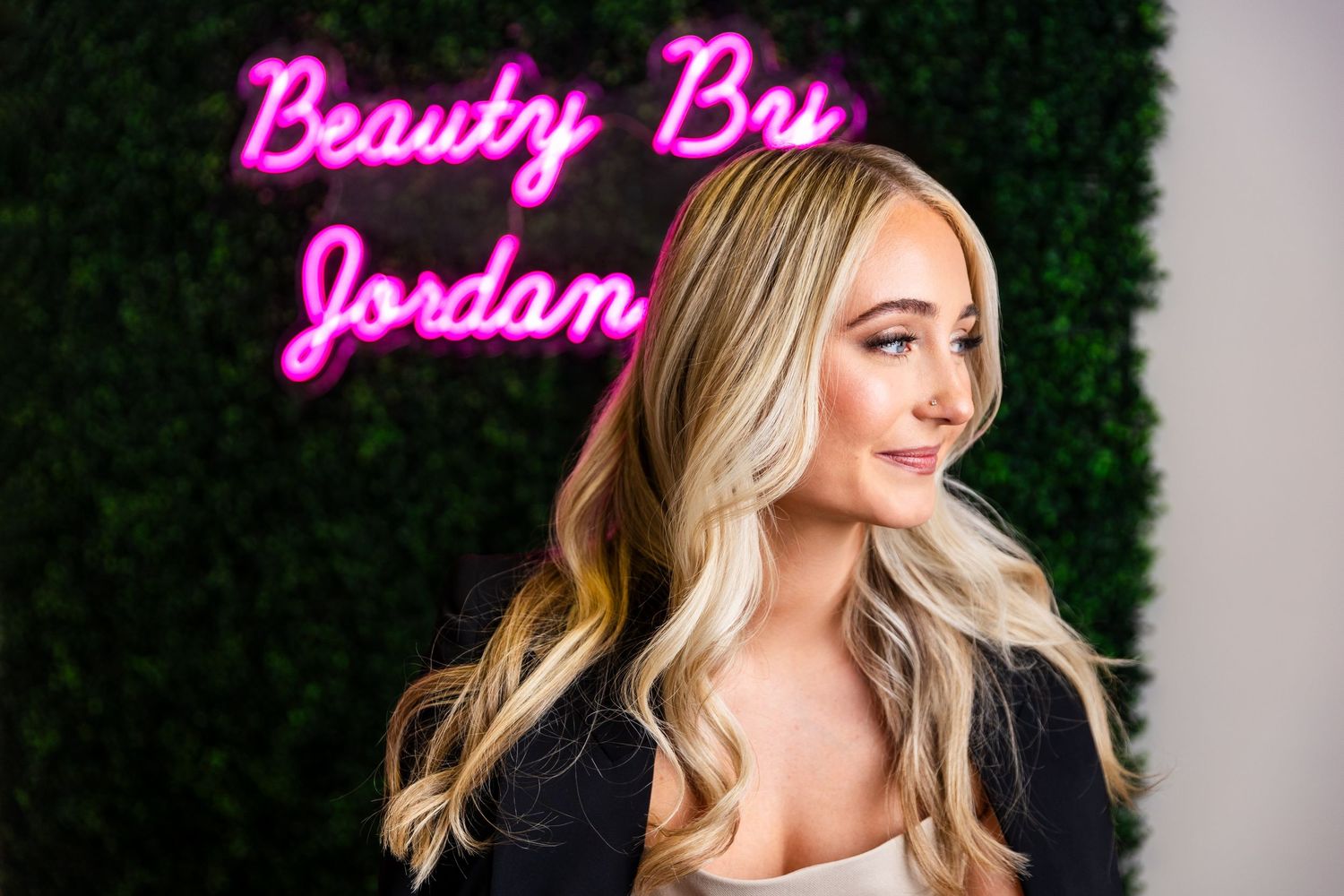 A woman with blonde wavy hair stands before a green hedge wall with pink neon 'Beauty Bar Jordan' signage.