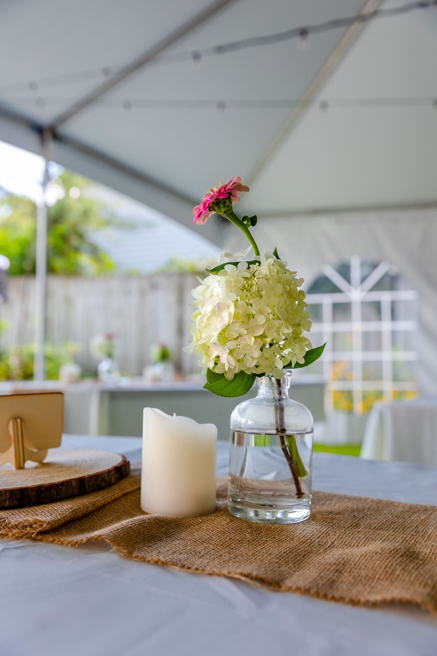 Candle and white hydrangea centerpieces on burlap runners create an elegant rustic table setting under a white tent.
