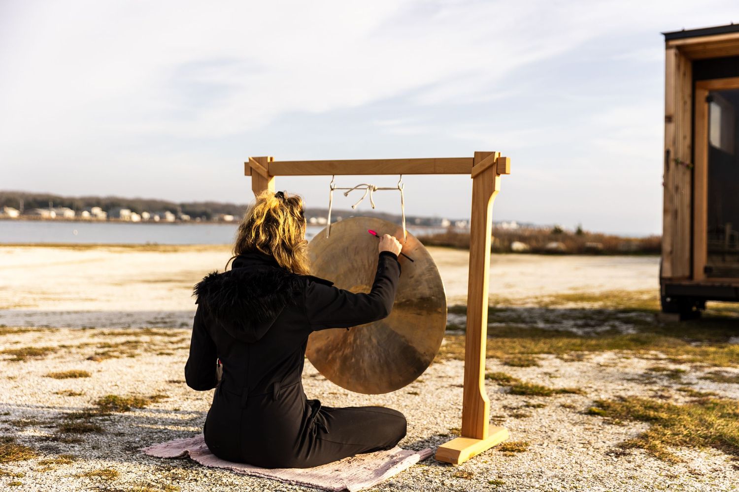 Person in black sits on snowy ground while striking large gong on wooden frame at beach during sunset.