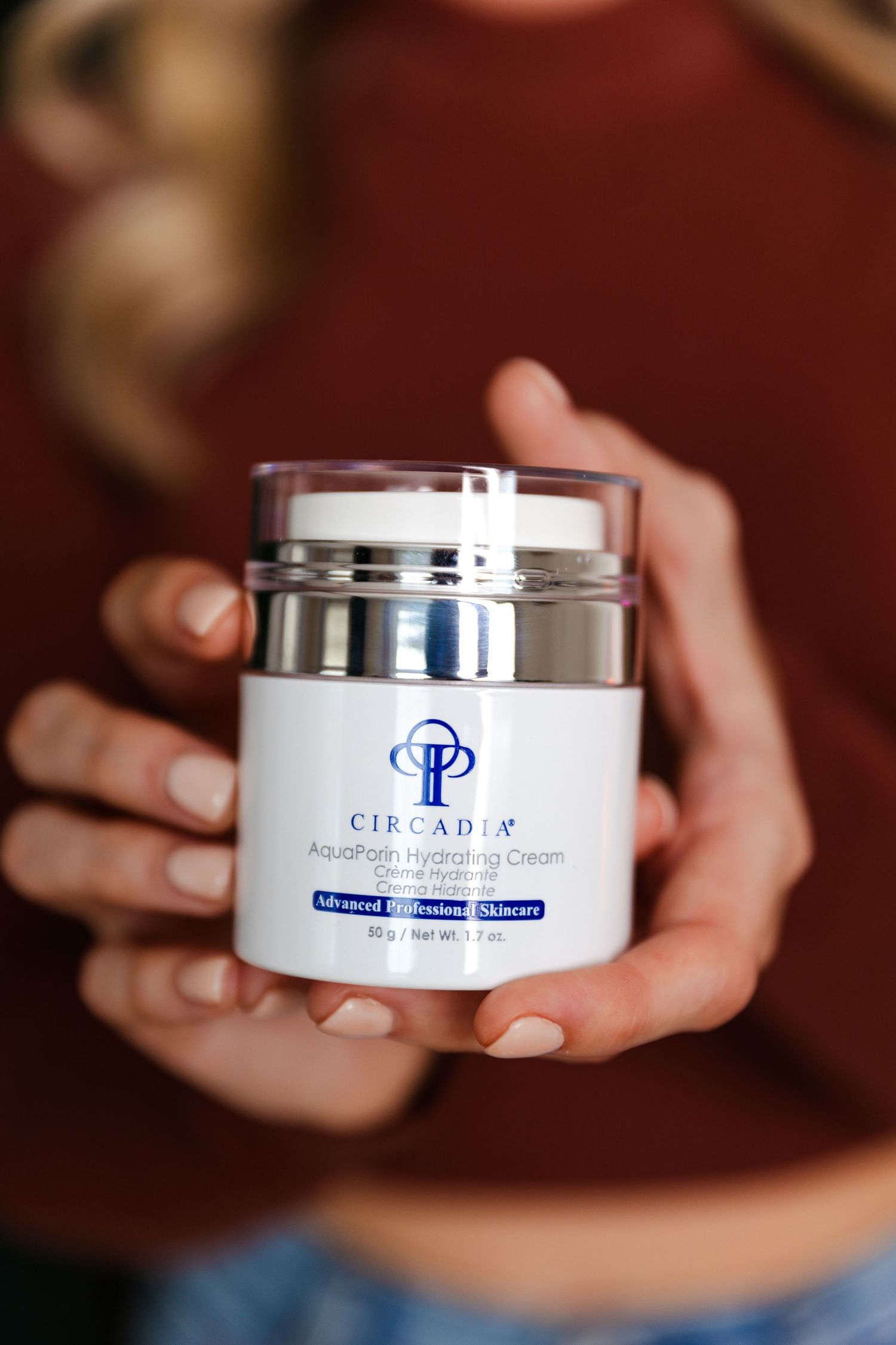 Hands holding a white cosmetic cream jar with blue CP logo against a blurred reddish background.
