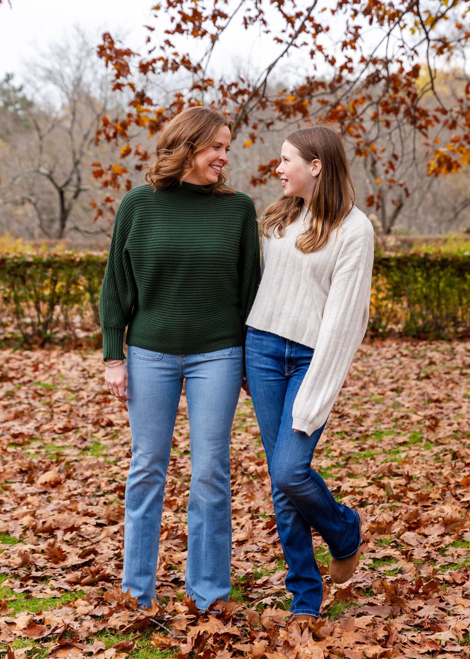 Fall scenery with two people in jeans and sweaters walking through colorful autumn leaves on the ground.