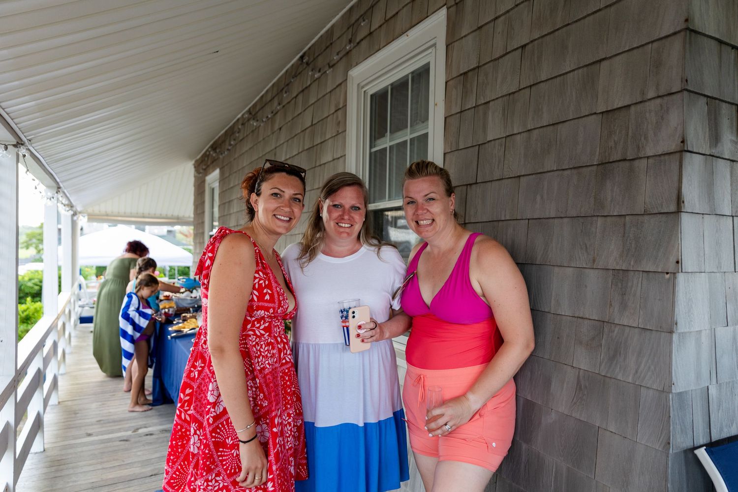 Three people in summer attire smile together on a covered porch beside a gray shingled building.