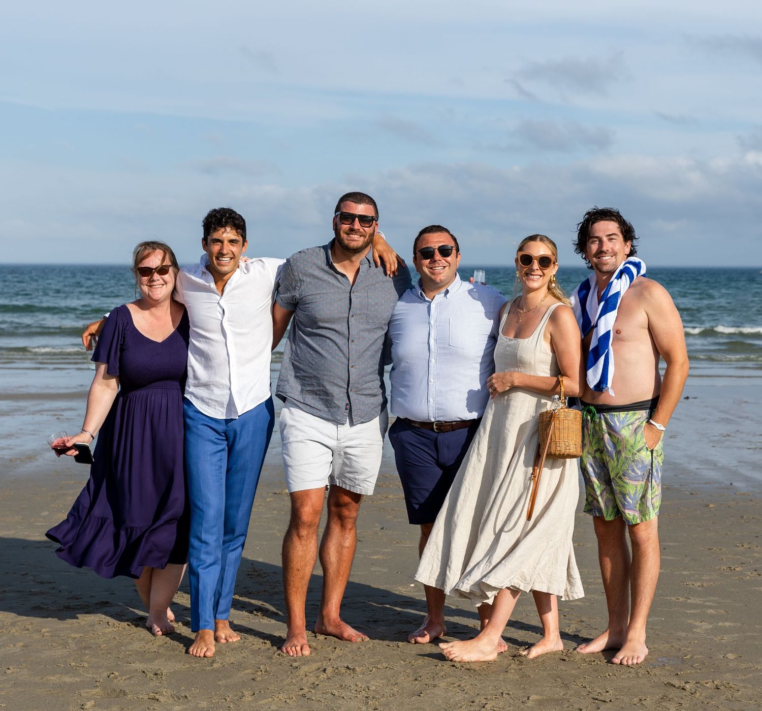 A group of friends stand together smiling on a beach with ocean waves and cloudy sky in the background.