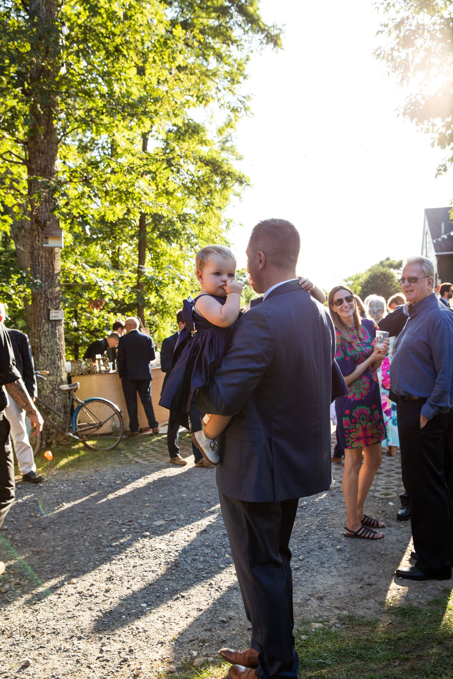 Guests gather outdoors under sunlit trees during an event as soft golden light filters through the branches.
