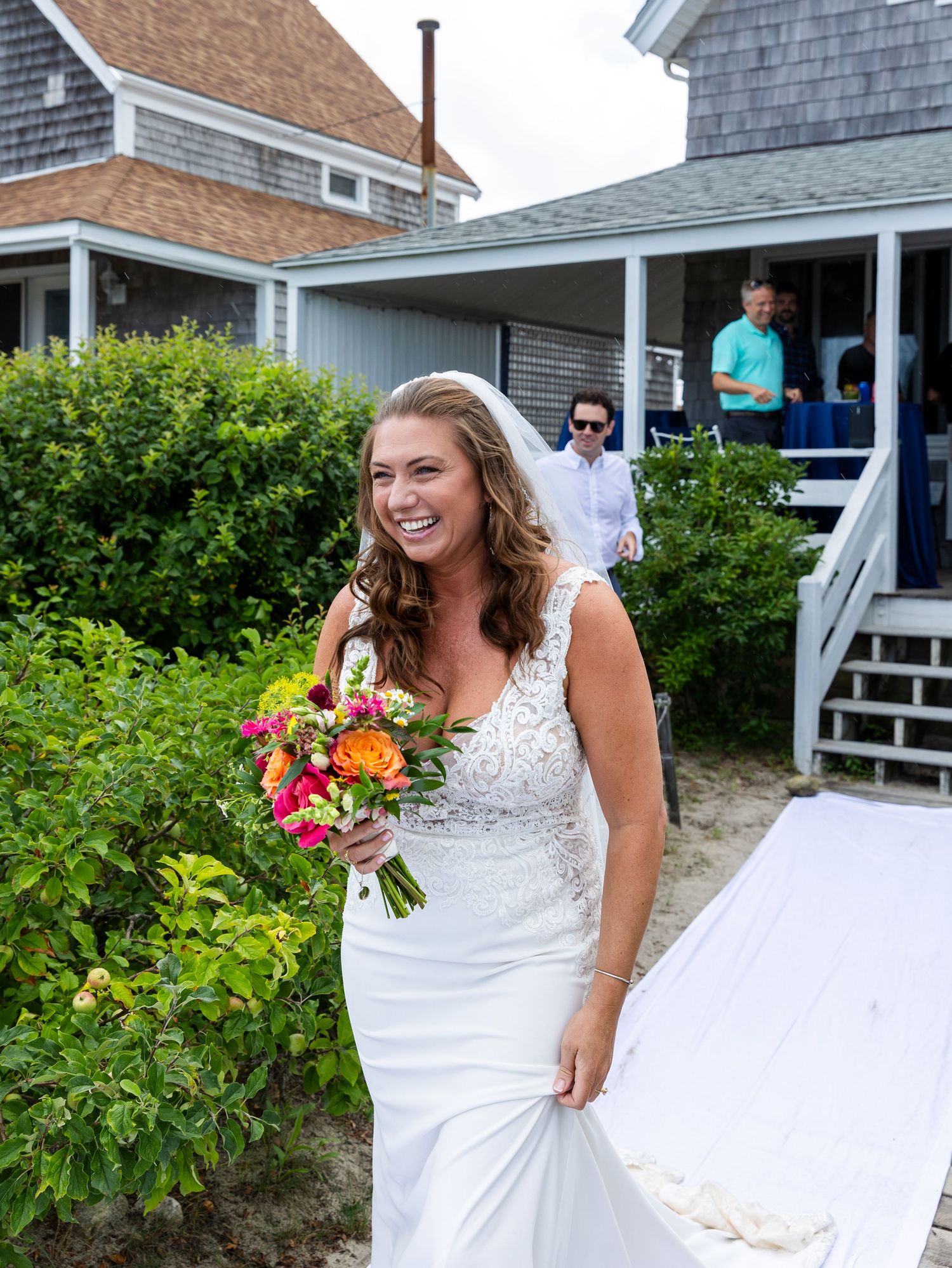 A bride in a white lace dress holds a vibrant pink and orange bouquet while standing next to lush greenery.
