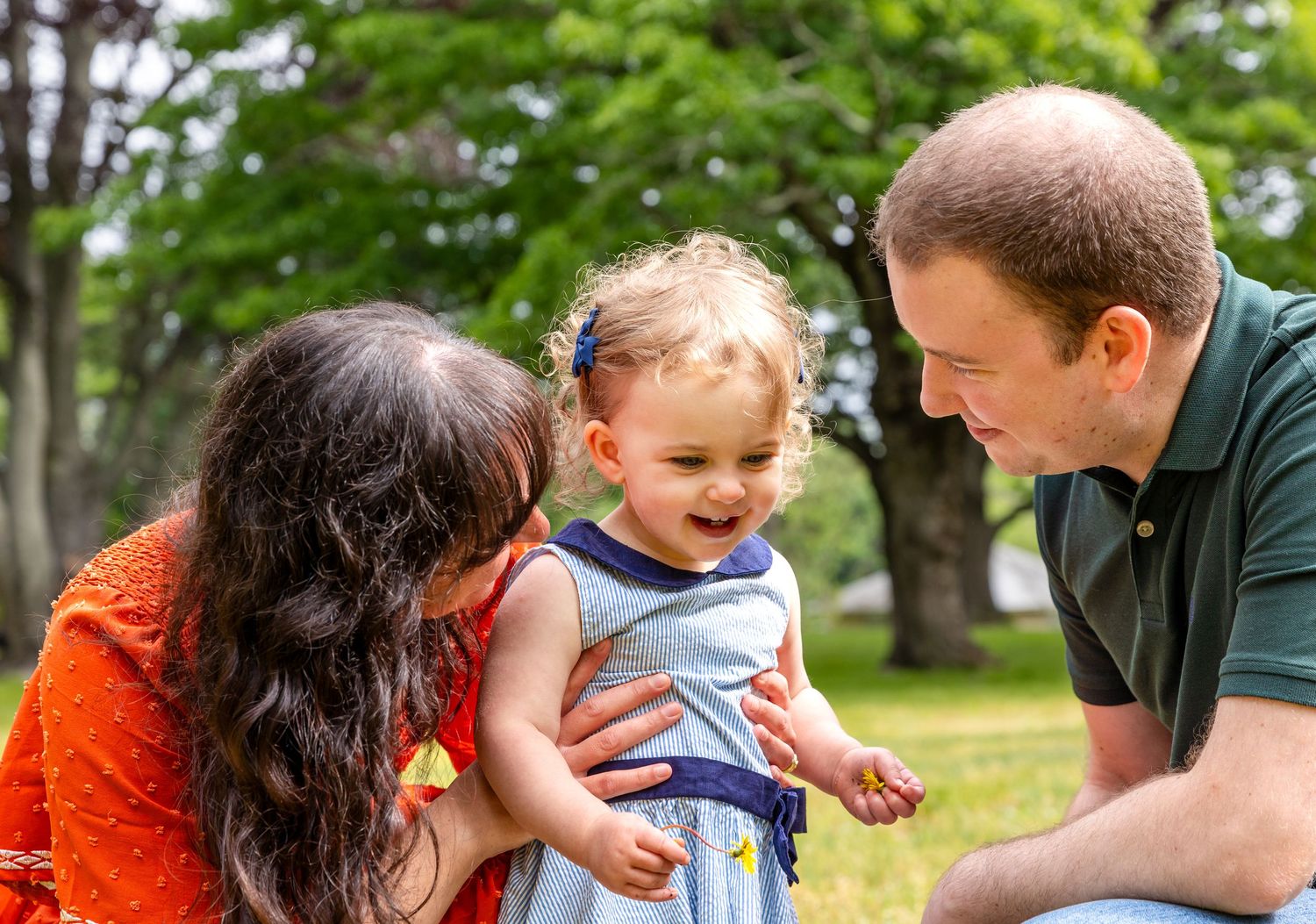 A joyful family moment outdoors with a laughing toddler in a blue outfit enjoying time with parents in a park setting.