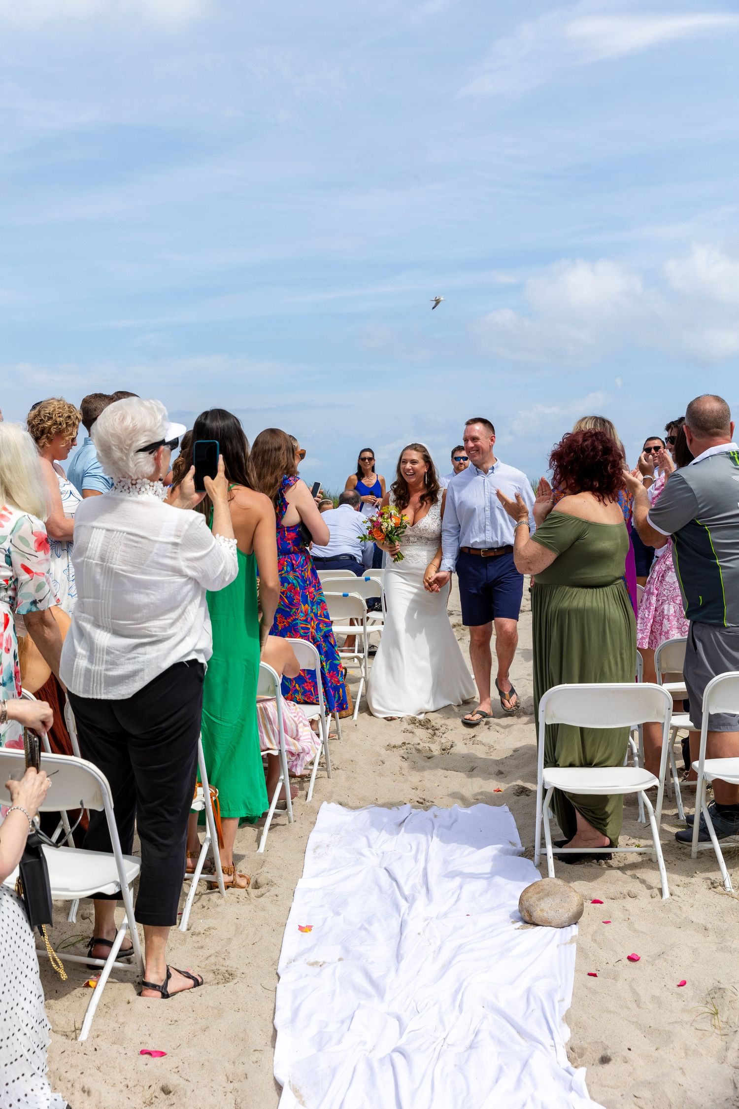 A beachside wedding ceremony setup with a white aisle runner and folding chairs as guests gather on the sand.