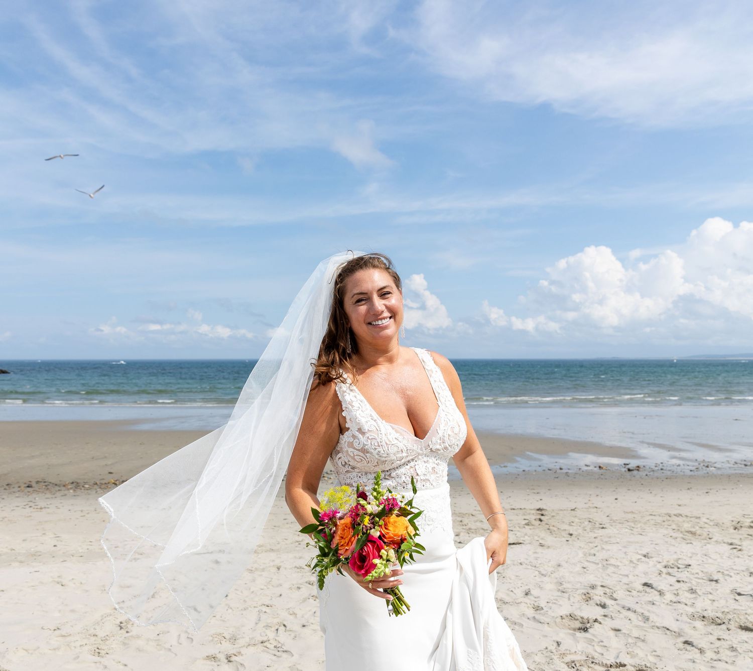 A bride in a white wedding dress holds a vibrant bouquet while standing on a sunny beach with ocean waves and blue sky.