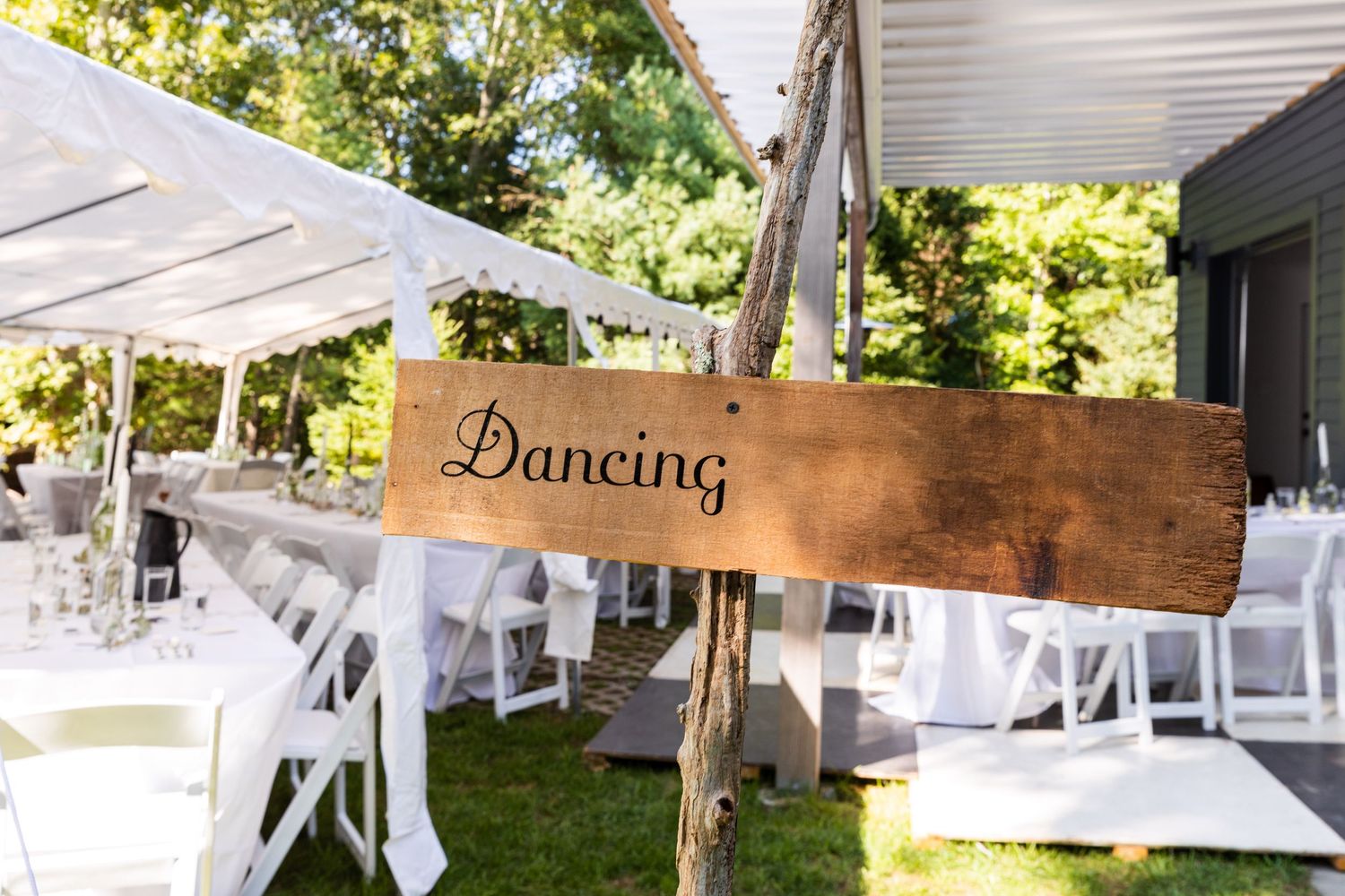 Wooden directional sign reading 'Dancing' hangs over outdoor wedding reception setup with white folding chairs.