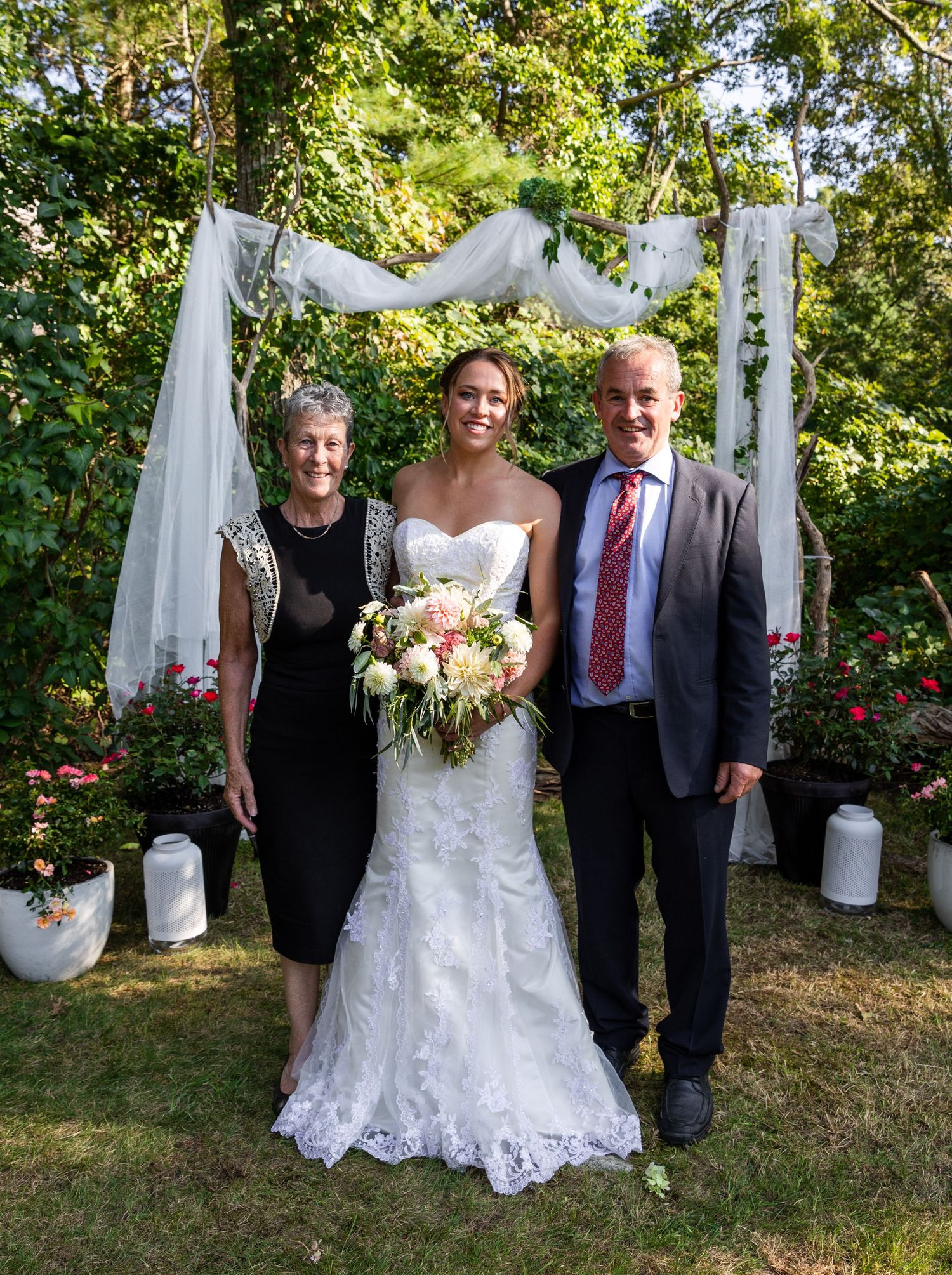 Wedding ceremony scene in garden with white draped arch, floral decorations and happy couple posing outdoors.