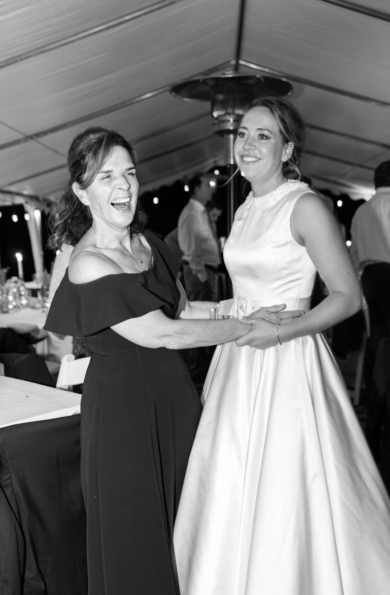 Two people share joyful embraces and dance moments at an indoor reception under a white tent.