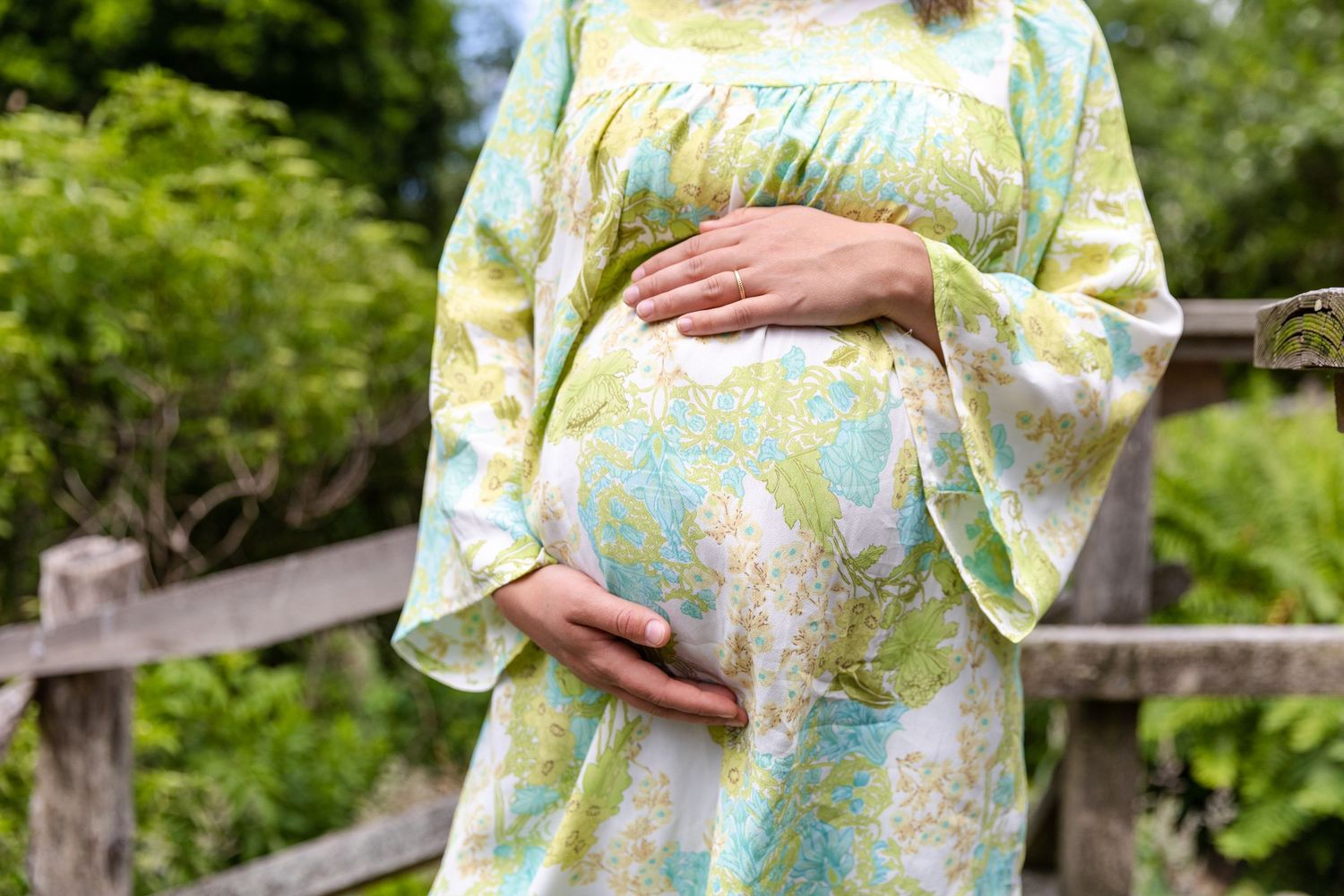 A pregnant person wearing a floral kimono-style dress cradles their baby bump while standing outdoors in a garden setting.