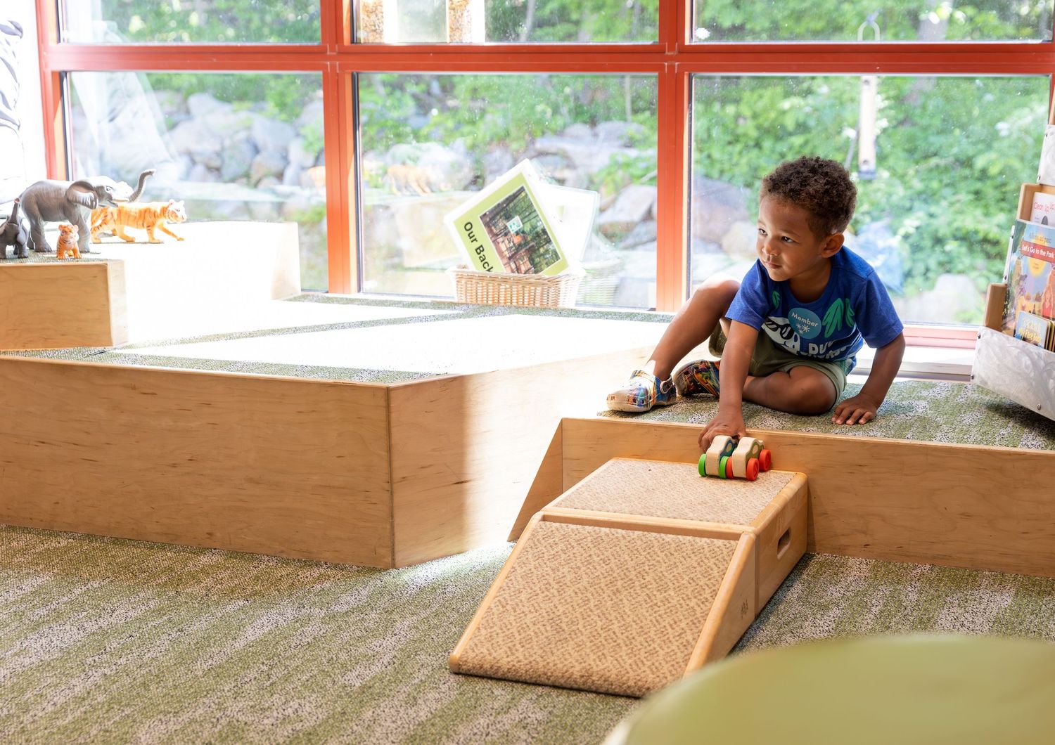 Young child playing with a toy car on a wooden ramp setup in a sunny room with large windows.
