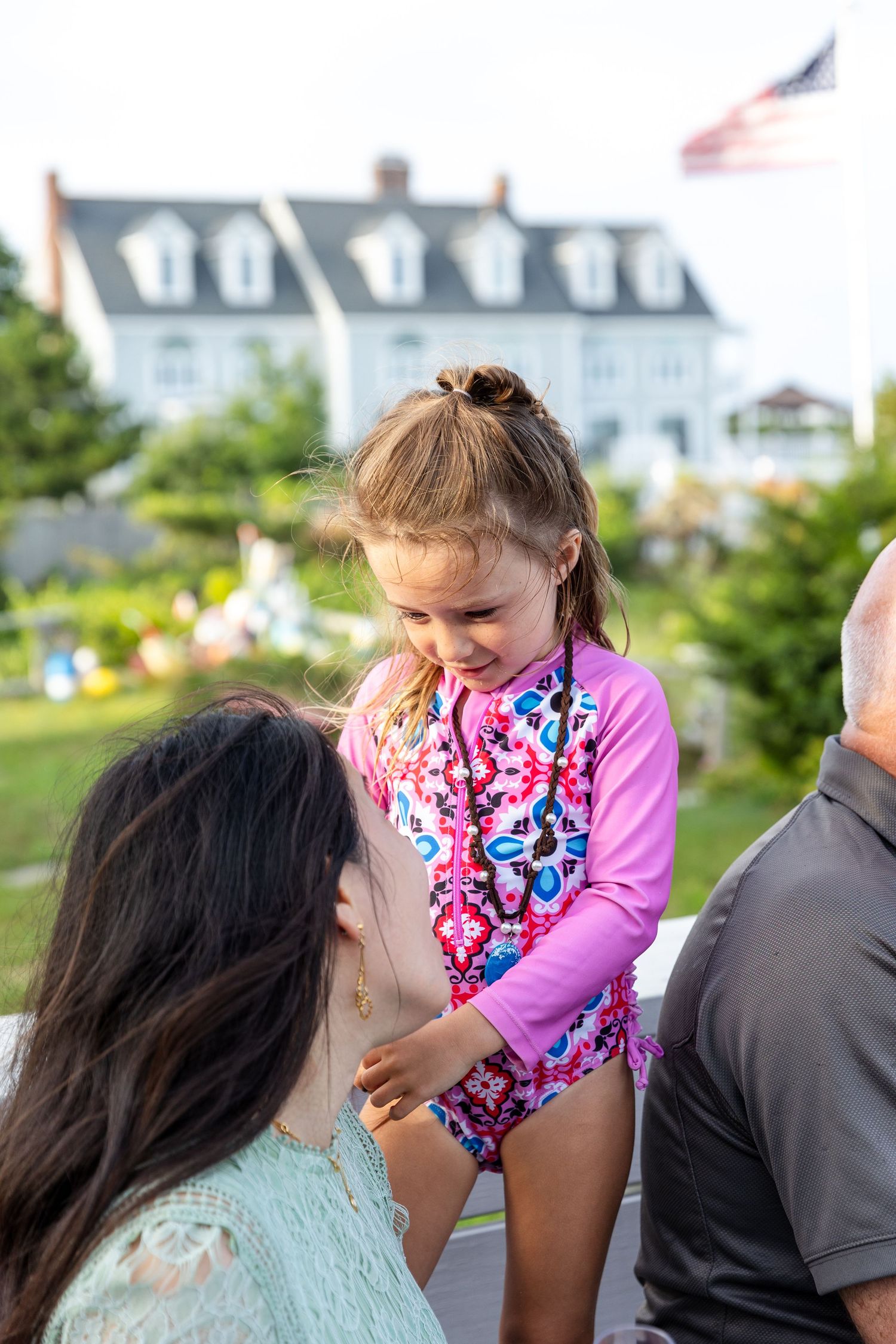 A child in a colorful floral swimsuit plays outdoors with a classic New England colonial house in the background.
