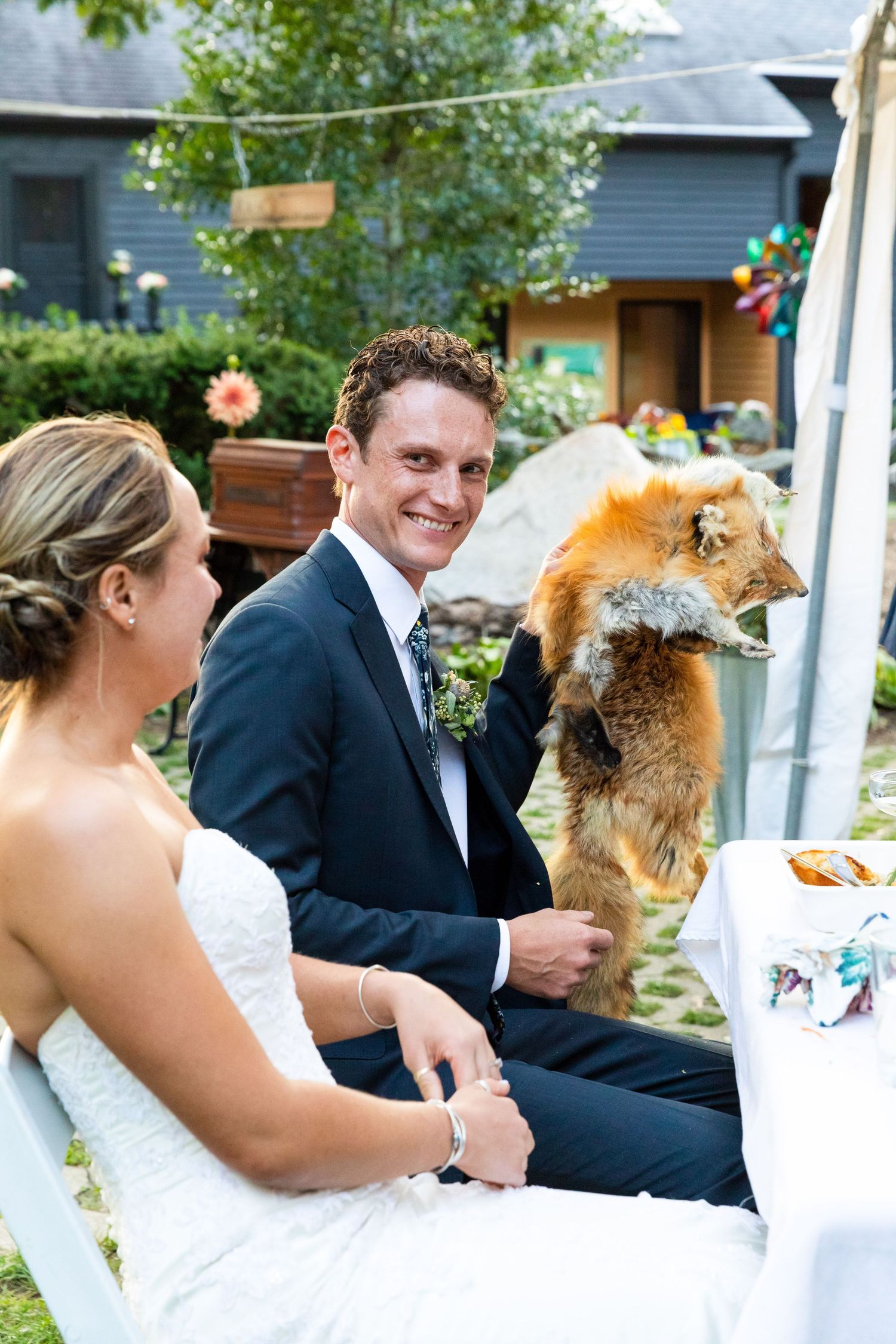 A wedding couple sitting outdoors getting photobombed by their smiling red-furred dog.