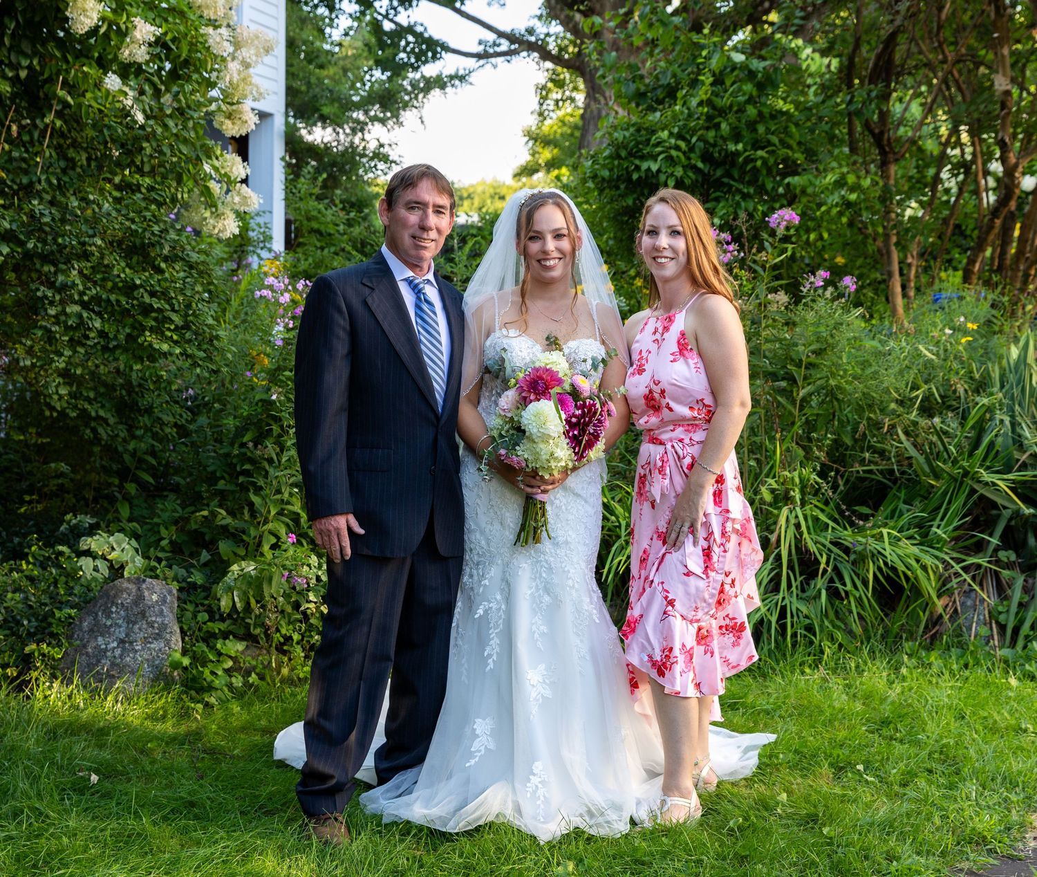 Three people pose together in a lush garden setting during a wedding celebration with greenery and flowers.