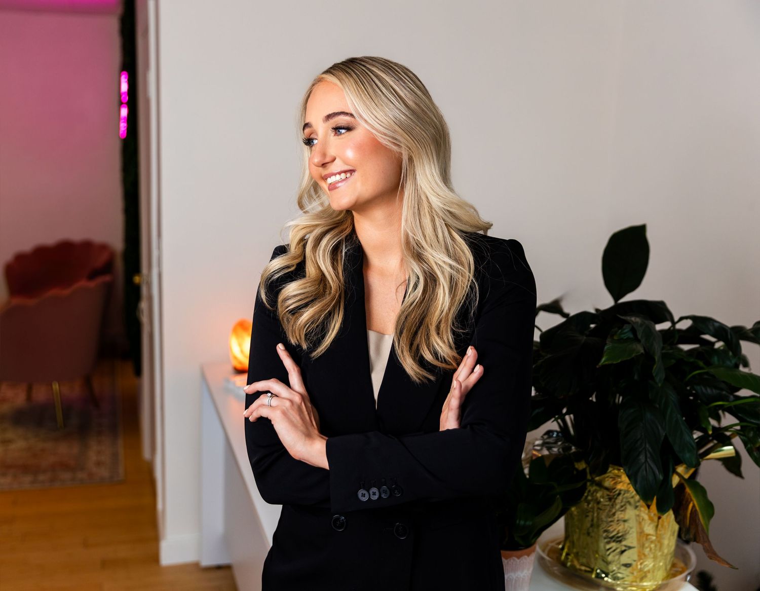 A woman in a black top sits smiling in front of a desk with a phone, potted plant and window behind her.