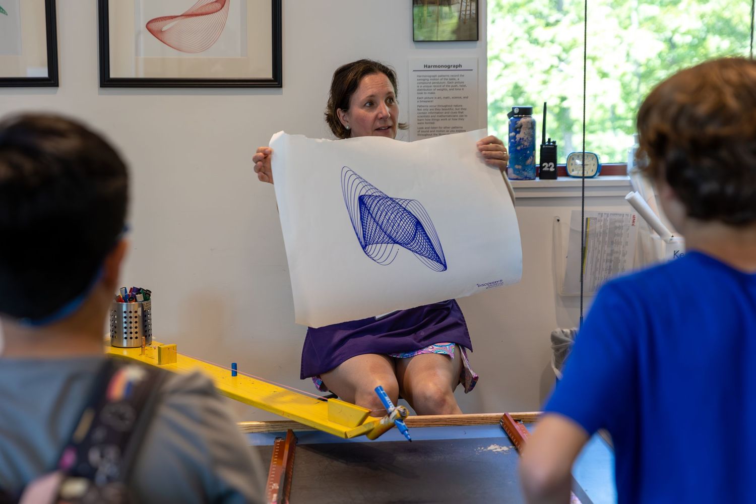 A person works on a drawing at a desk with white paper while others sit nearby in an office setting.