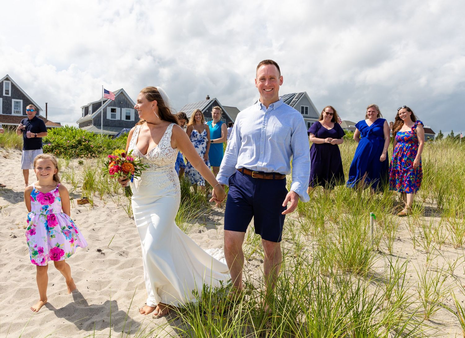 A procession of wedding guests walk along a scenic coastal beach path surrounded by tall grass and dunes during sunset.