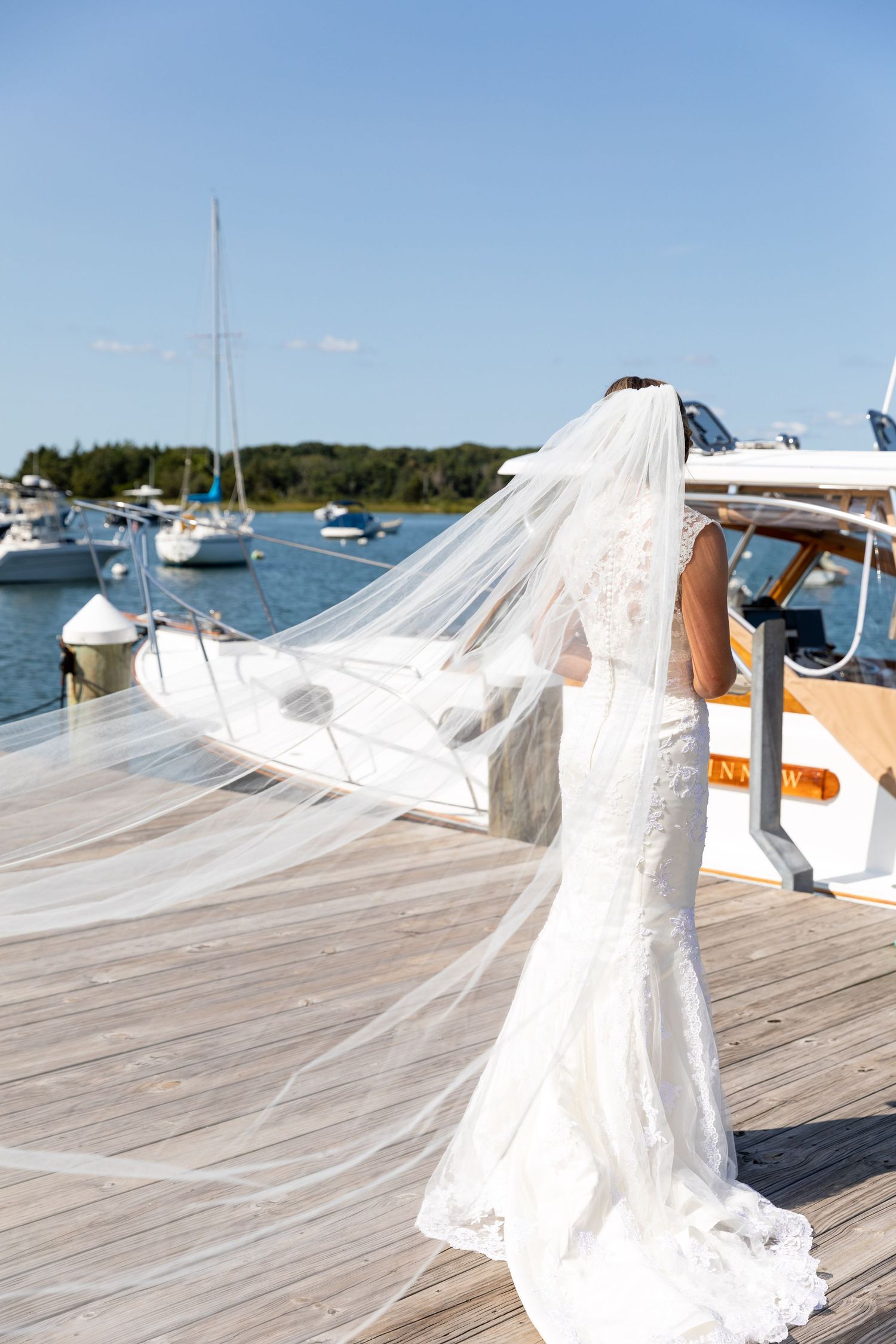 A flowing white wedding dress and veil trailing in the wind on a wooden dock with boats in the marina background.