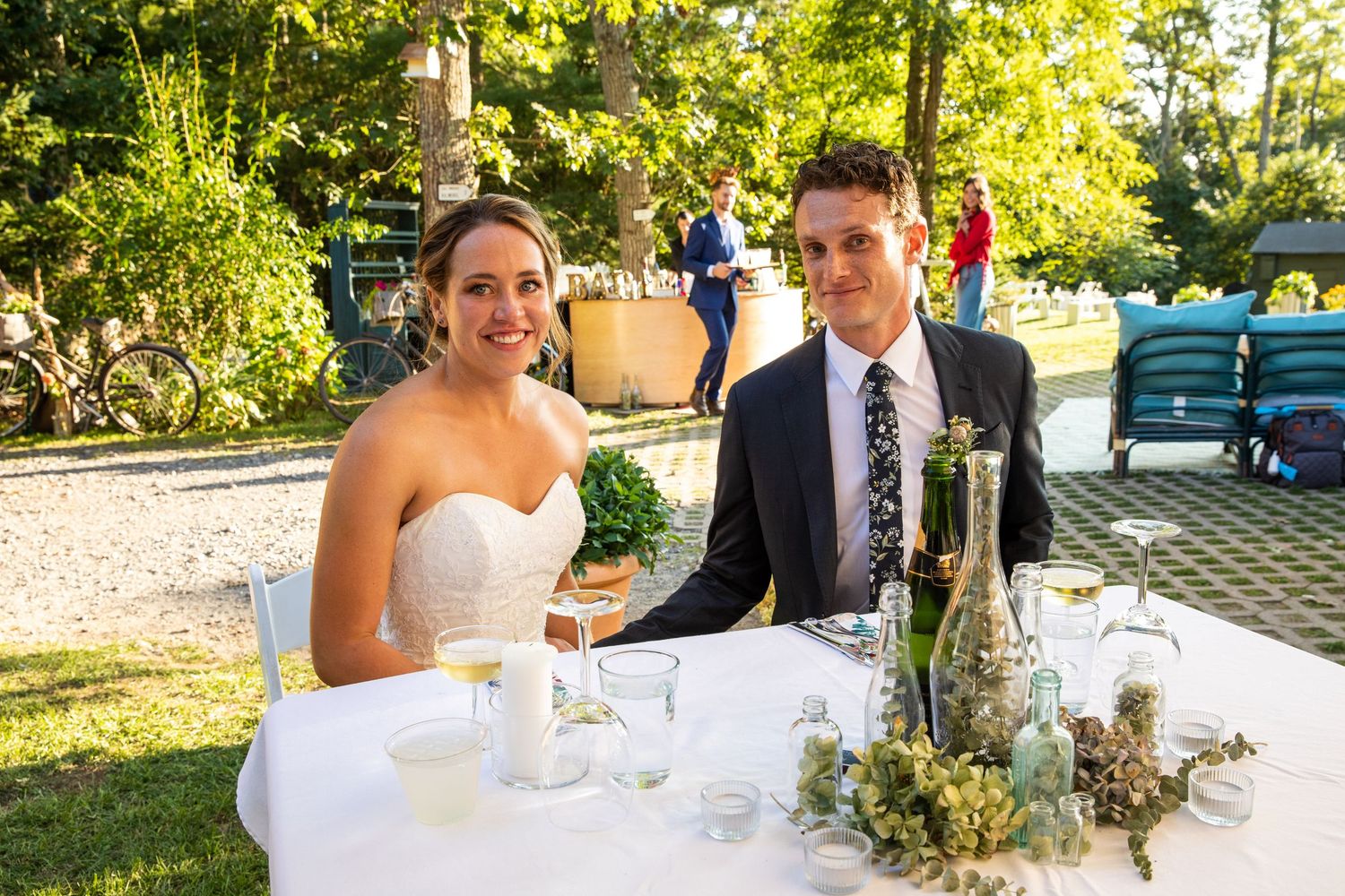 A couple sits at an elegant outdoor reception table with wine glasses and floral centerpieces in a sunny garden setting.