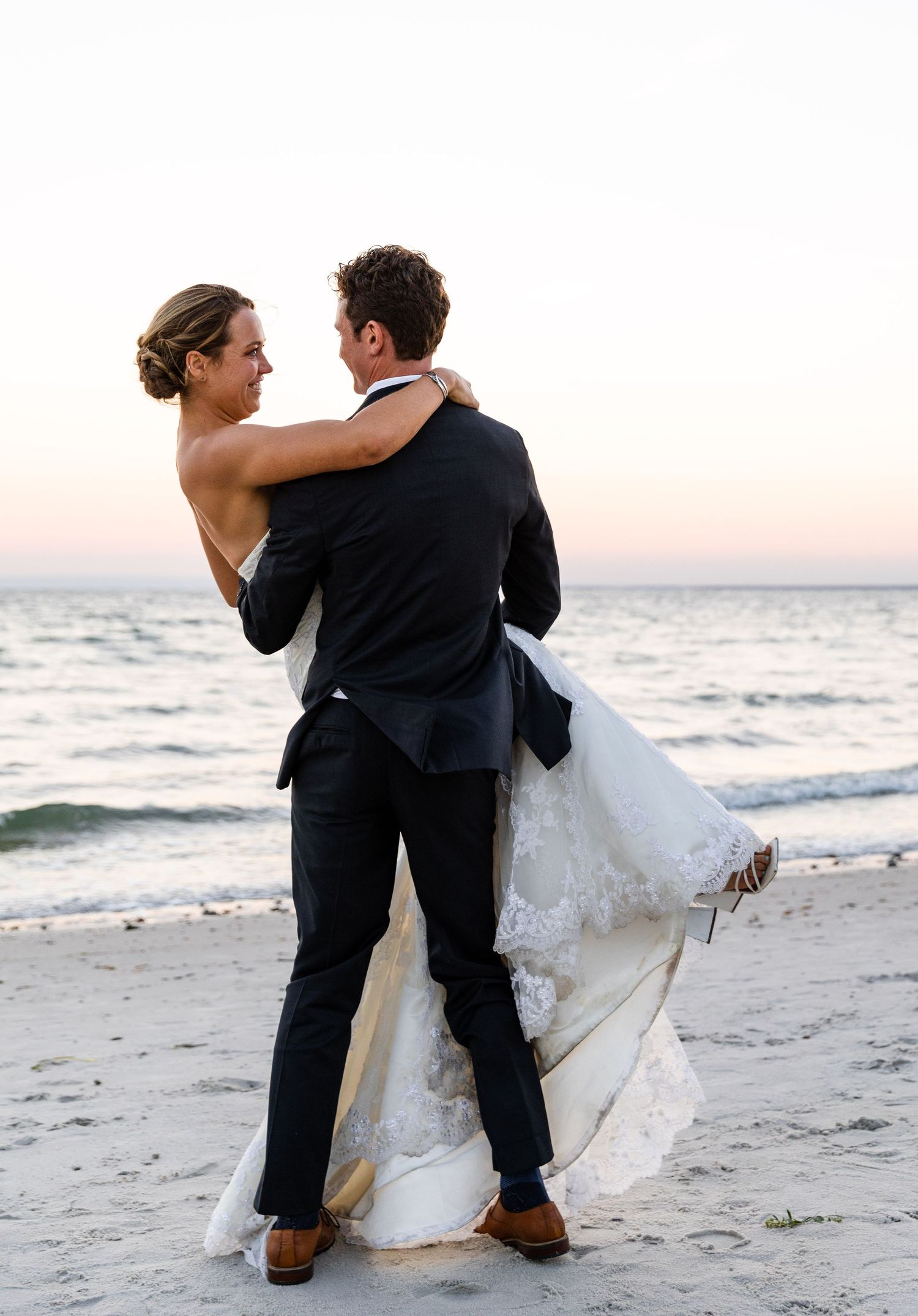A couple in wedding attire dances intimately on a beach at sunset while waves roll in behind them.