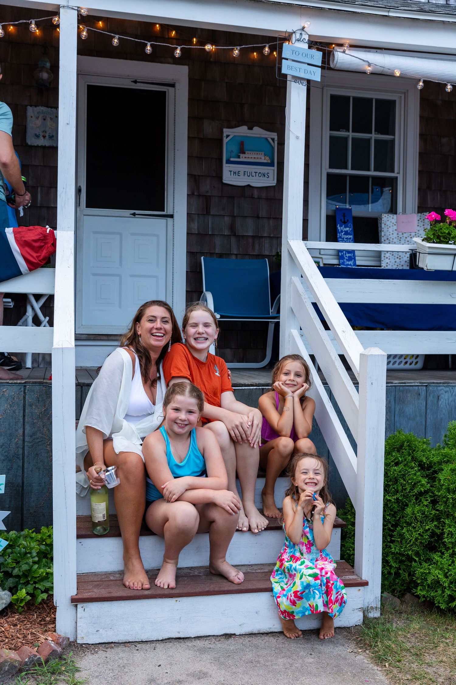 Group of people sitting on a white porch with hanging string lights and greenery visible in the summer setting.