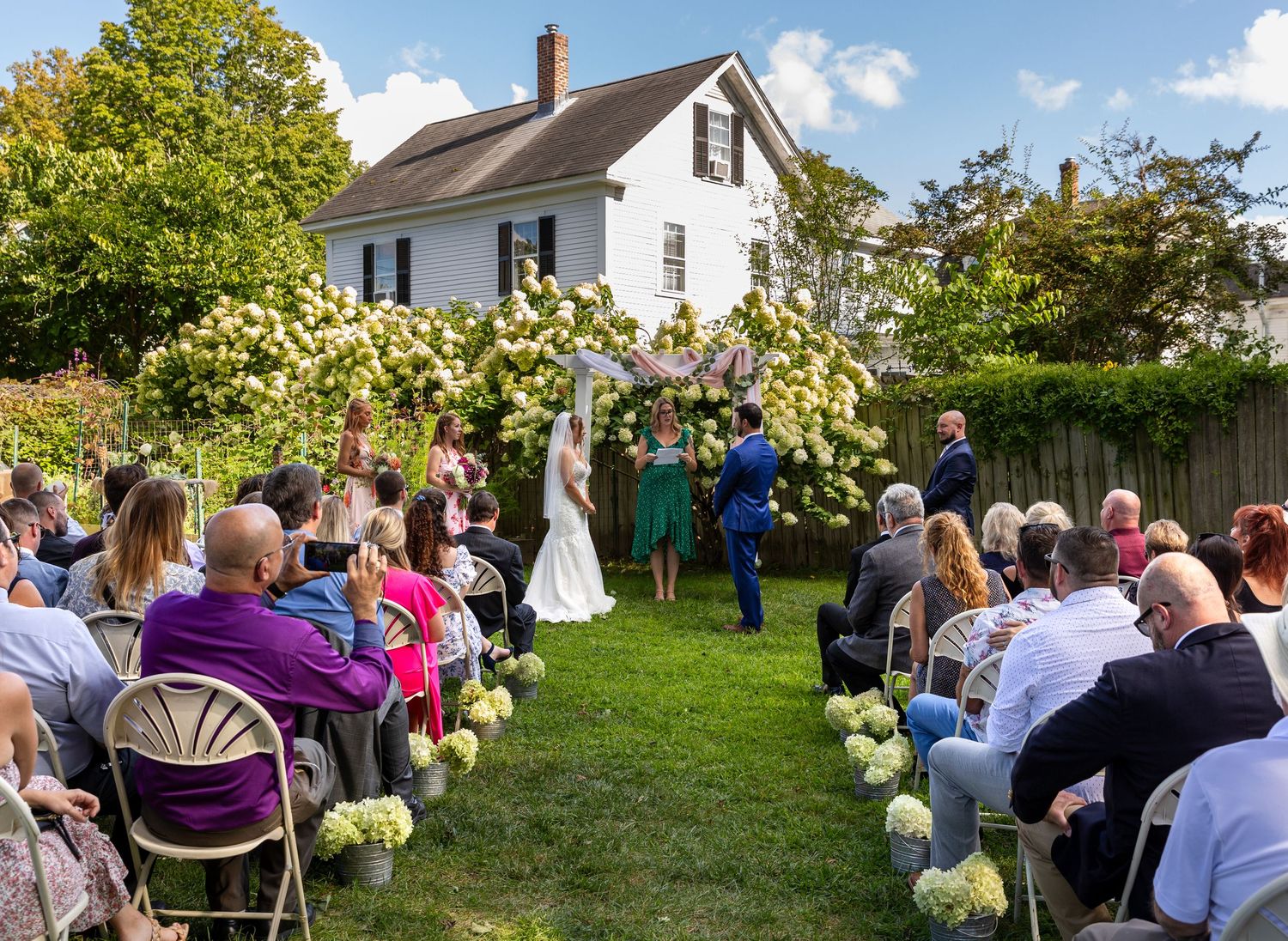 Wedding guests gather in a sunny garden for an outdoor ceremony surrounded by green trees and white farmhouse buildings.